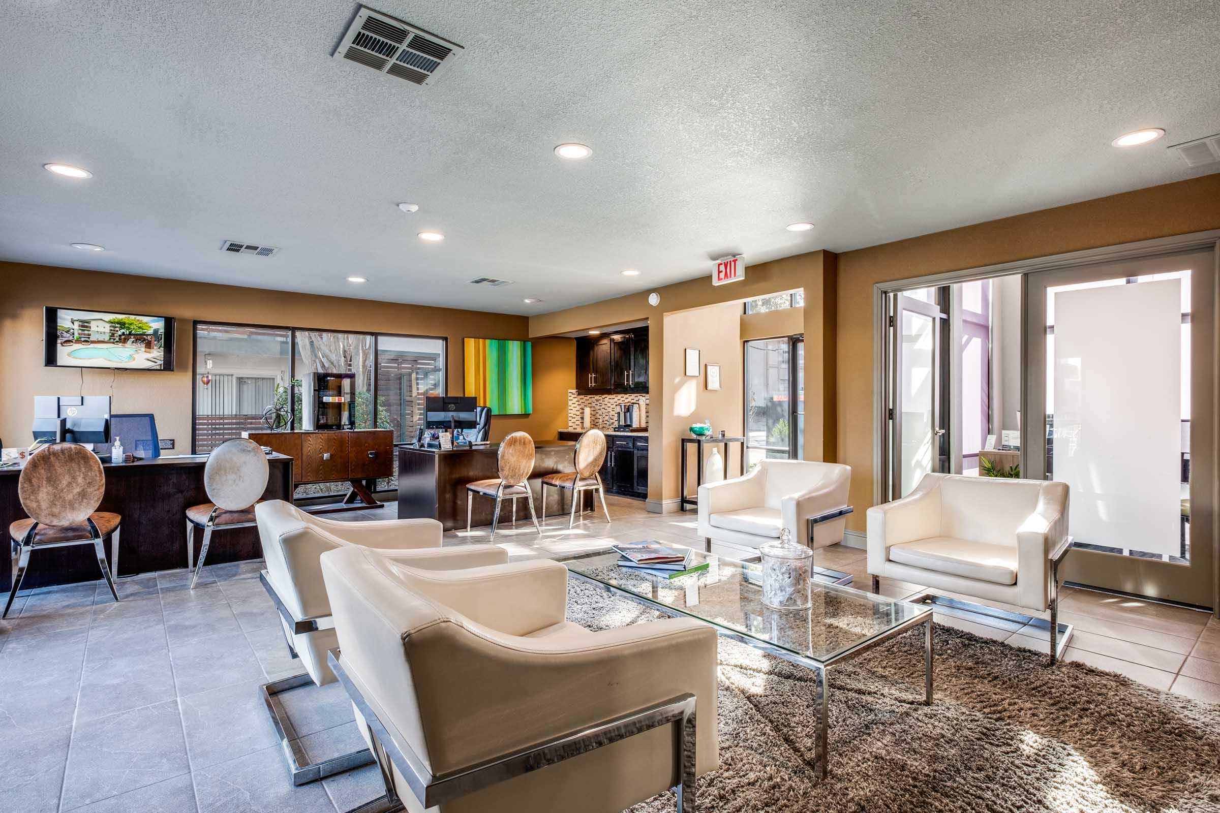 Modern lobby of a building featuring sleek white chairs, a glass coffee table on a plush rug, and a front desk with a dark wooden finish. The space is well-lit with natural and artificial light, showcasing a welcoming atmosphere. Two televisions and decorative elements add to the contemporary design.