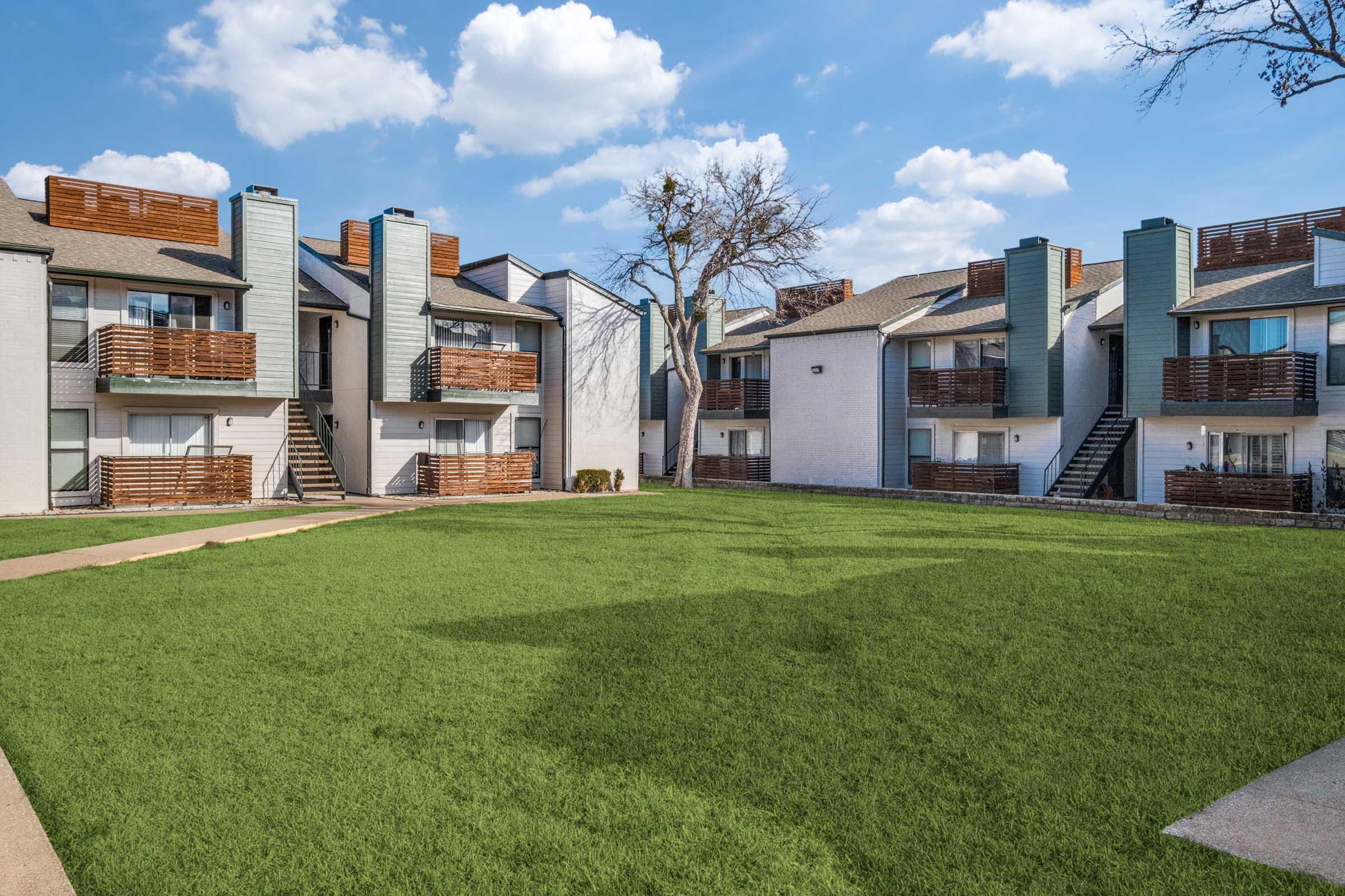 A view of a modern apartment complex featuring two-story buildings with wooden balconies. The central area is a well-maintained green lawn, surrounded by walkways. The sky is partly cloudy, creating a bright and inviting atmosphere.