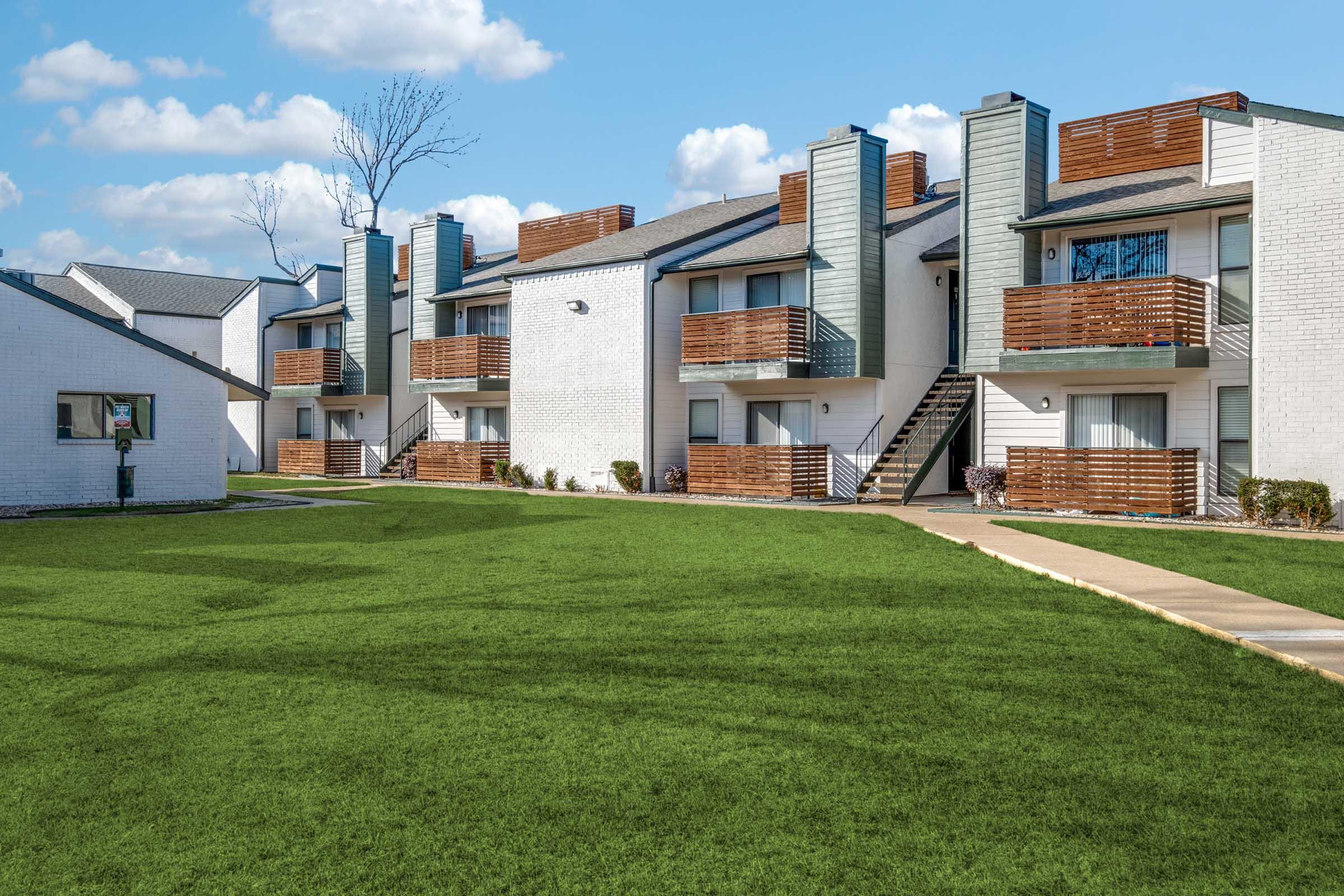 A sunny day in a residential area featuring modern apartment buildings with wooden balconies. The buildings are spaced around a green lawn, with a sidewalk leading through the area. A few trees and scattered shrubs add to the landscape, creating a welcoming community atmosphere.