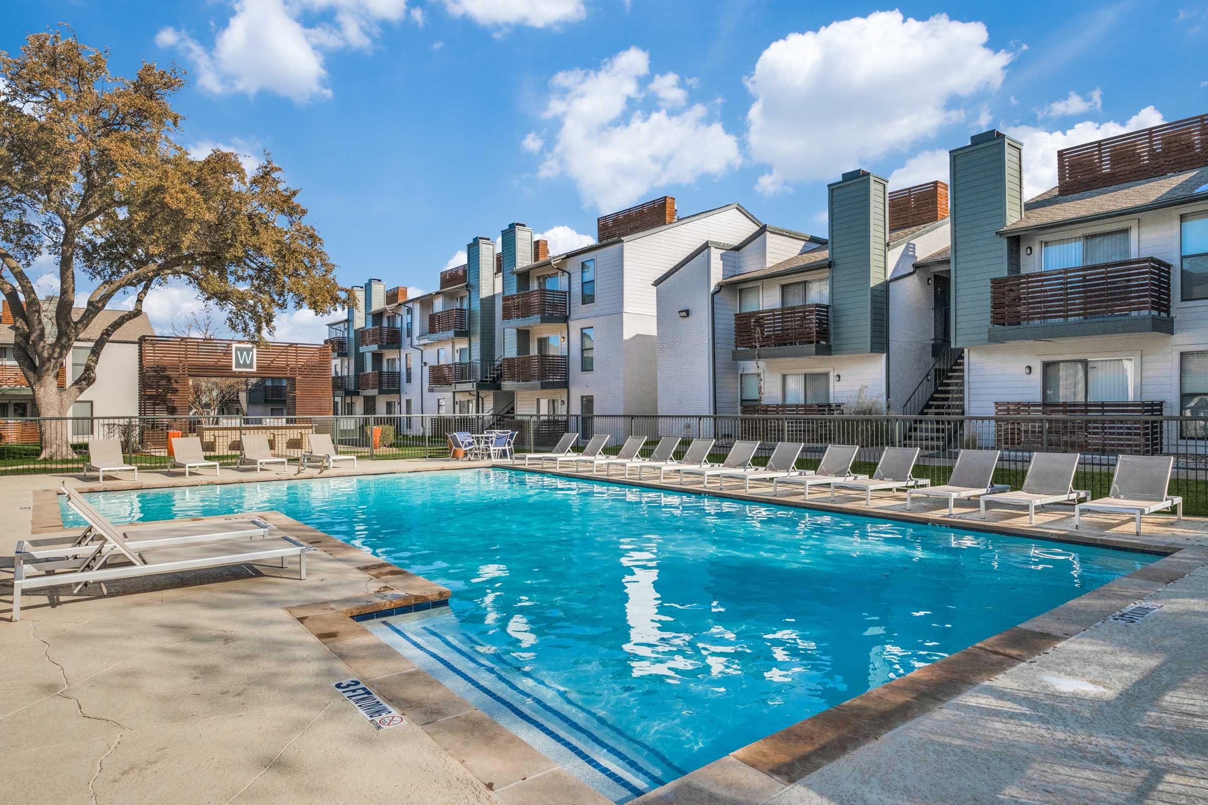 A residential swimming pool with lounge chairs, surrounded by modern apartment buildings. The clear blue water reflects the sky, and a few trees provide shade nearby. Sunny weather with scattered clouds enhances the inviting atmosphere of the pool area.