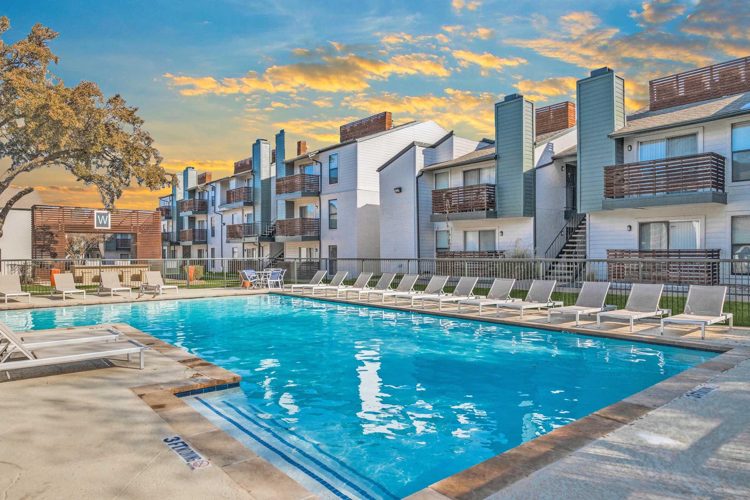 A clear blue swimming pool surrounded by lounge chairs, with an apartment complex in the background. The scene features green trees and a vibrant sunset sky, creating a relaxing atmosphere.