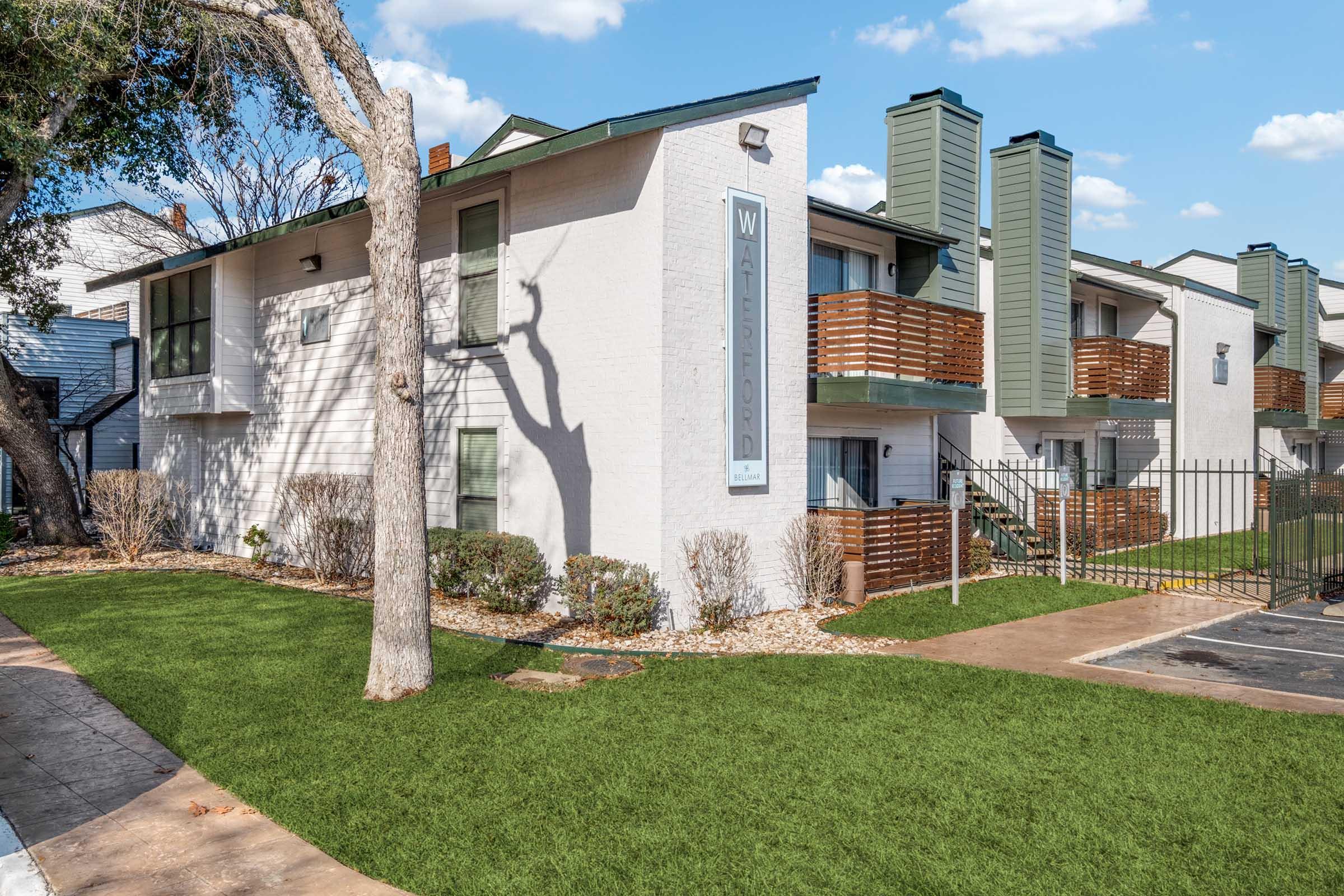Modern apartment complex exterior featuring light-colored brick walls, green accents, and a well-manicured lawn. The building has multiple balconies and is surrounded by trees and shrubs. A gated entry leads to a parking area, with a clear blue sky and fluffy clouds in the background.