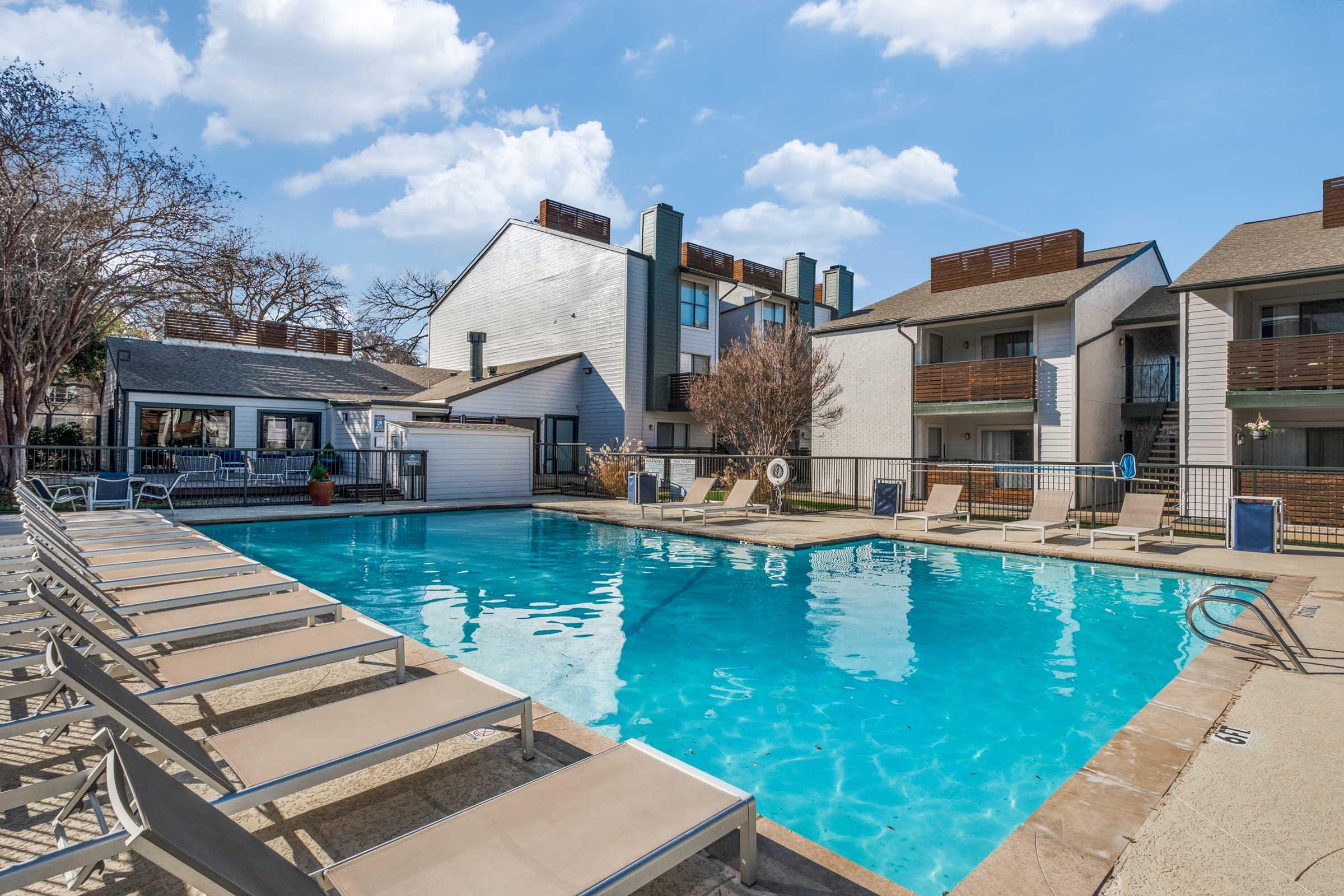 A clear swimming pool surrounded by lounge chairs, with residential buildings in the background and trees nearby. The sky is bright blue with a few fluffy clouds, creating a sunny, inviting atmosphere.