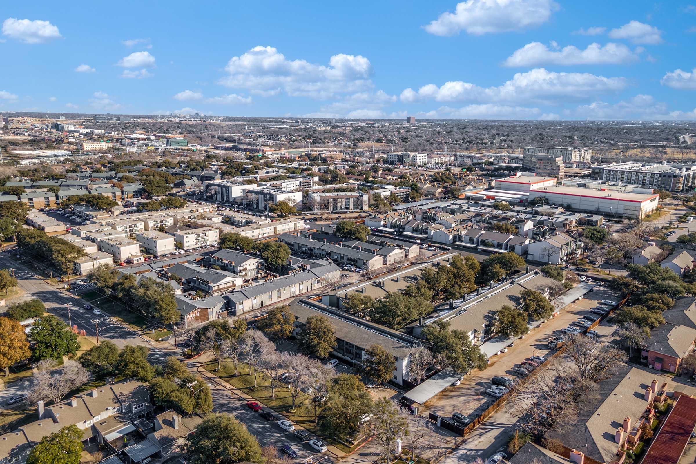 Aerial view of a suburban area featuring residential buildings, trees, and parking spaces. The landscape includes various structures and roads, with a clear blue sky and scattered clouds in the background, showcasing a mix of urban and natural elements.