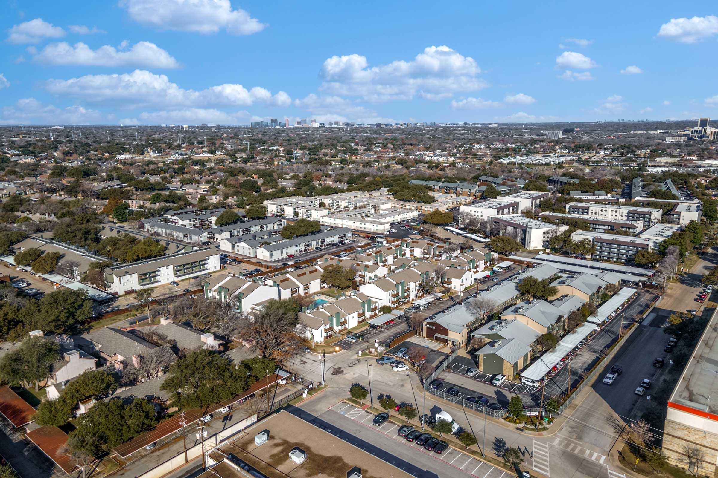 Aerial view of a suburban neighborhood featuring a mix of residential buildings, including townhouses and low-rise apartments, surrounded by trees and open spaces. In the background, a city skyline can be faintly seen under a partly cloudy sky.