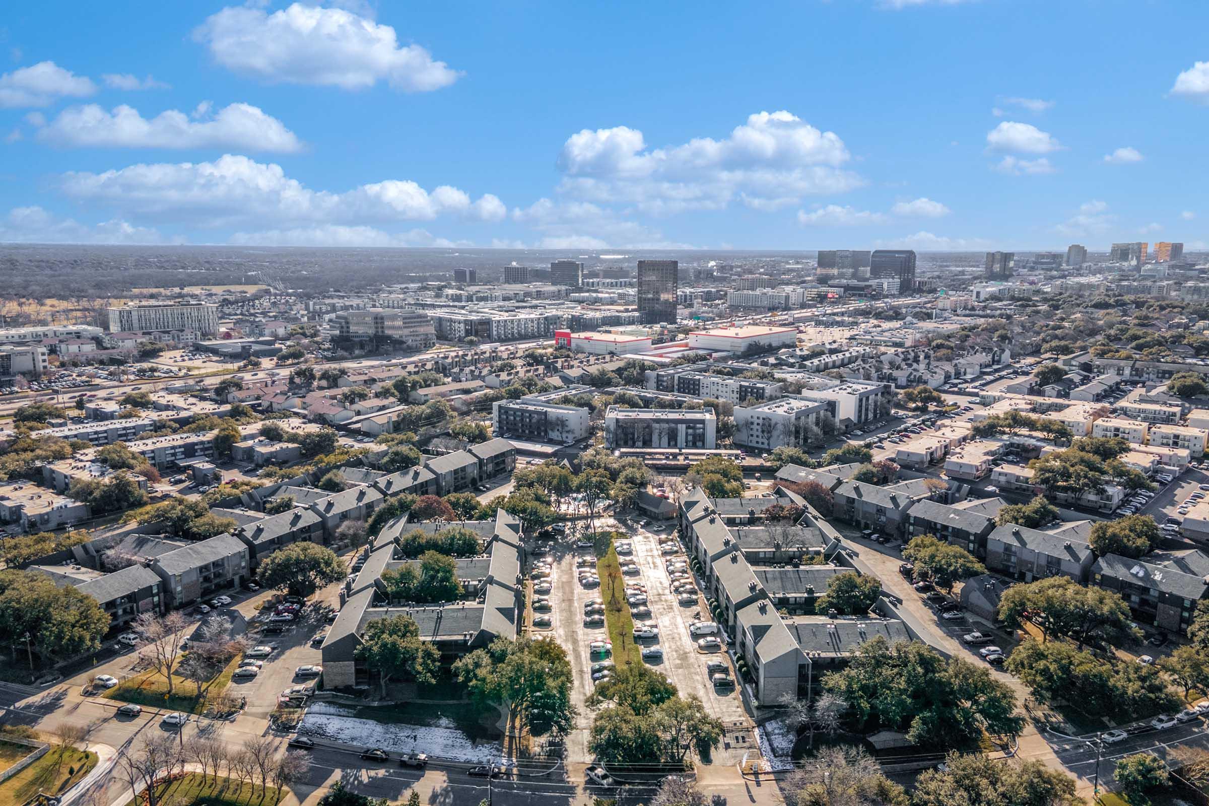 Aerial view of a suburban area featuring clustered townhouses surrounded by a parking lot and greenery. In the background, a skyline with various commercial buildings is visible under a blue sky with scattered clouds.