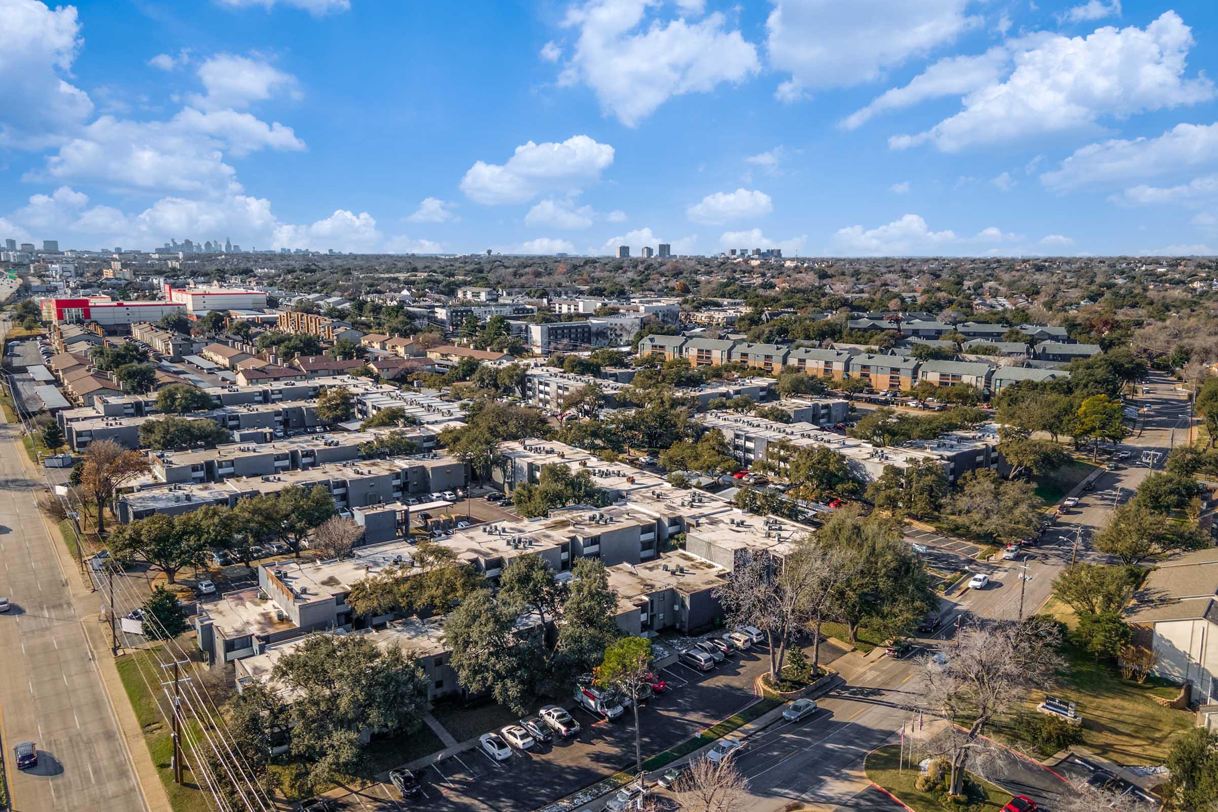 Aerial view of a suburban neighborhood featuring several low-rise apartment buildings, trees, and parking areas. In the background, a city skyline is visible under a bright blue sky with scattered clouds. The scene depicts a mix of residential and commercial areas.