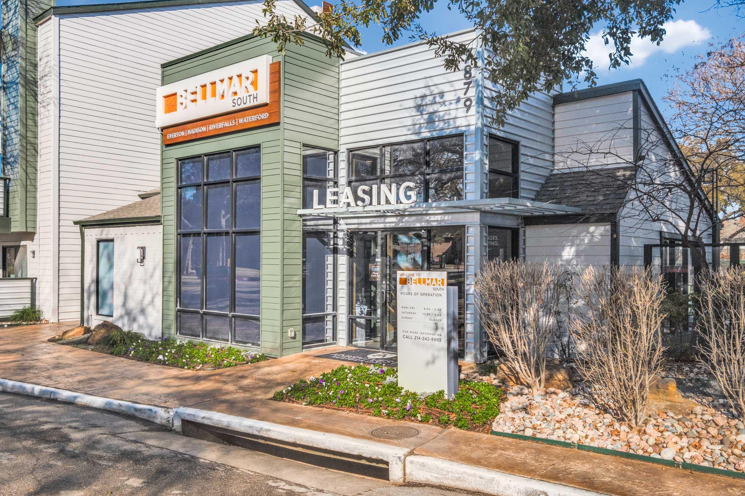 Leasing office of a modern residential complex named "Lumar South," featuring a contemporary architectural design. The building has large glass windows and a green exterior, surrounded by landscaped plants and a clear blue sky. A sign indicates leasing information, adding to the inviting atmosphere.