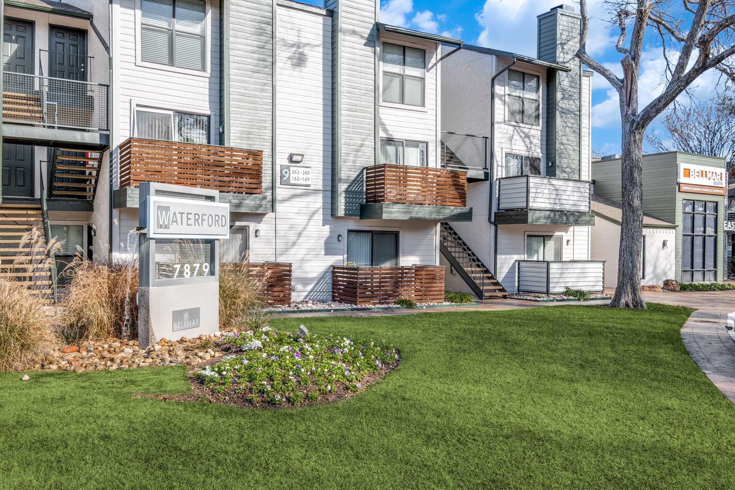 A well-maintained apartment complex with the sign "Waterford" in front. The building features modern architecture with balconies, surrounded by landscaped greenery and flowers. The sky is clear with a few clouds, and nearby structures are visible.