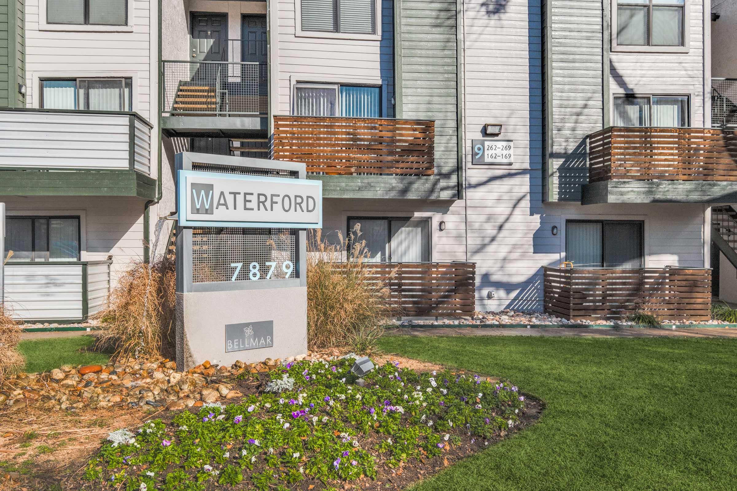 Sign for Waterford Apartments at 7879, featuring a modern multi-story building with green and gray exterior accents. Balconies with wooden railings are visible, along with landscaped areas including colorful flower beds and well-maintained grass.