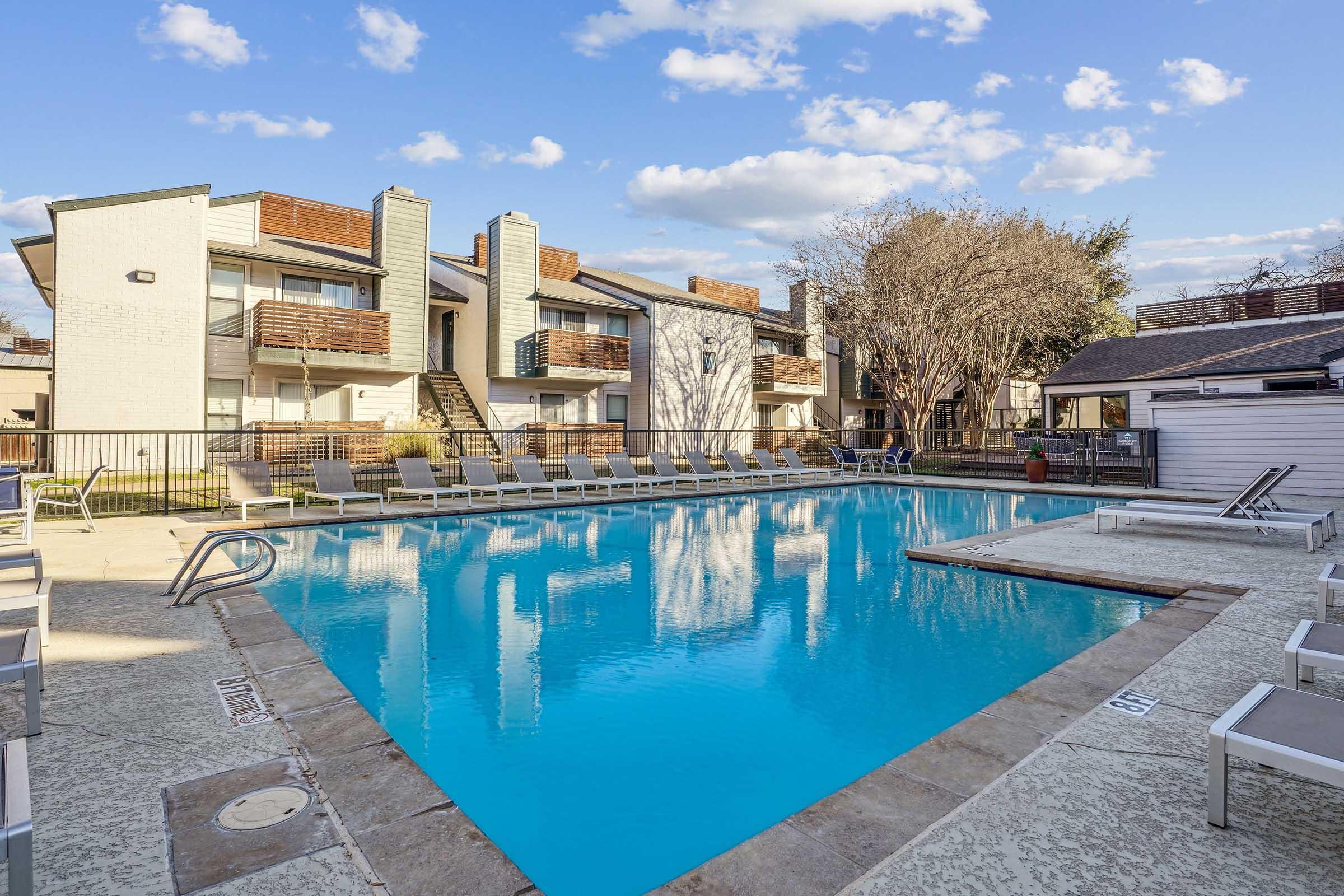 A welcoming apartment complex pool area featuring a large, clear blue swimming pool surrounded by lounge chairs. In the background, two-story buildings with balconies and trees are visible under a partly cloudy sky. The setting is inviting and ideal for relaxation.