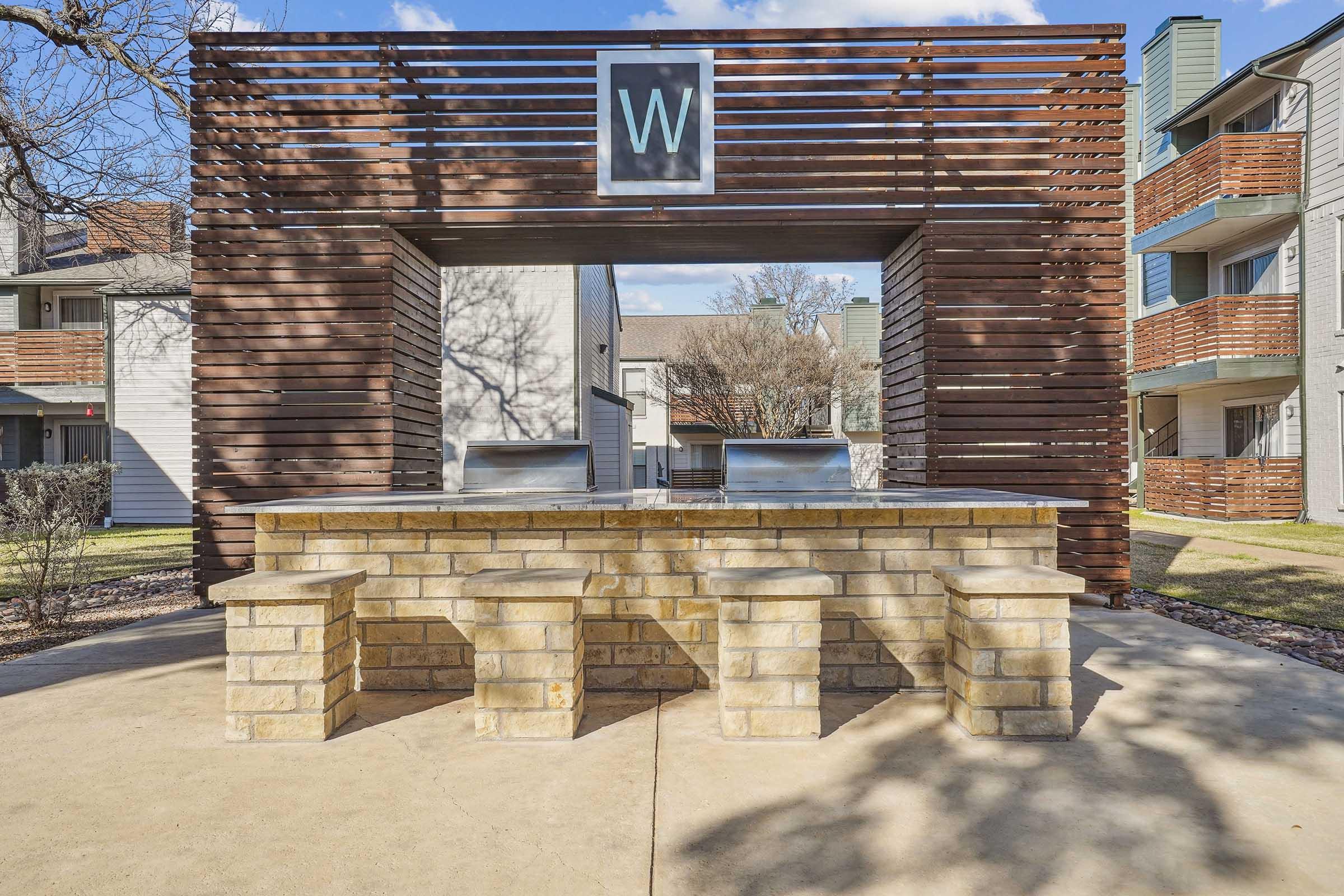 An outdoor communal grilling area featuring a stone table with integrated seating. The structure is made of wood slats and has a decorative panel with the letter "W." Nearby, there are trees and a grassy area, with apartments visible in the background. The sky is partly cloudy.