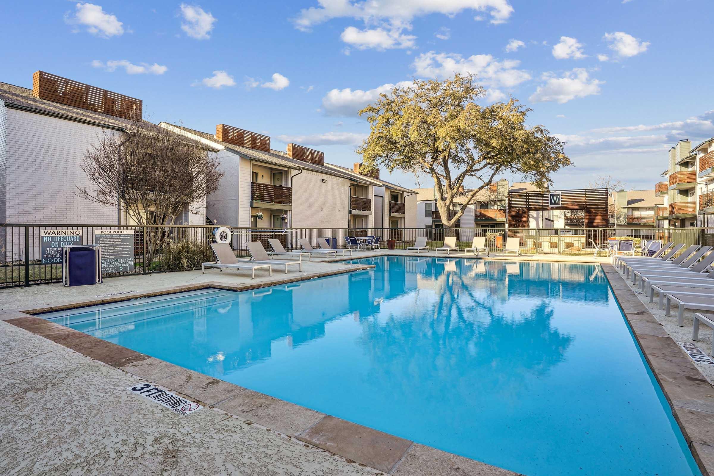 A clear blue swimming pool surrounded by lounge chairs, with an apartment building in the background. The sky is partly cloudy, and a large tree provides some shade near the pool area. The setting appears inviting and well-maintained, suitable for relaxation.