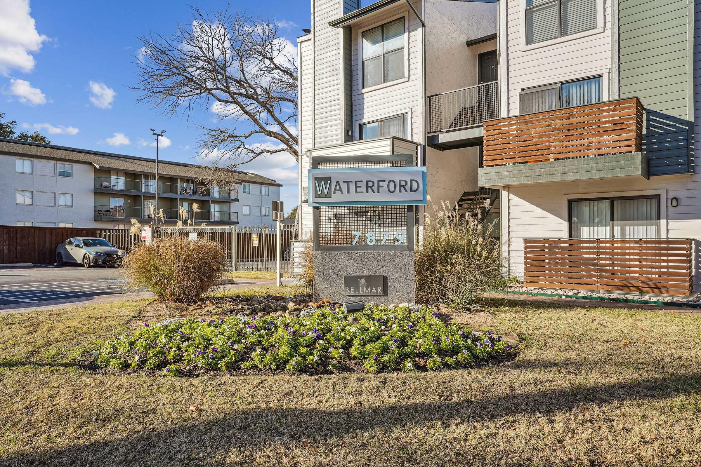 Sign for Waterford apartments at 7871, surrounded by landscaping with flowers. In the background, residential buildings and parking area are visible under a partly cloudy sky.