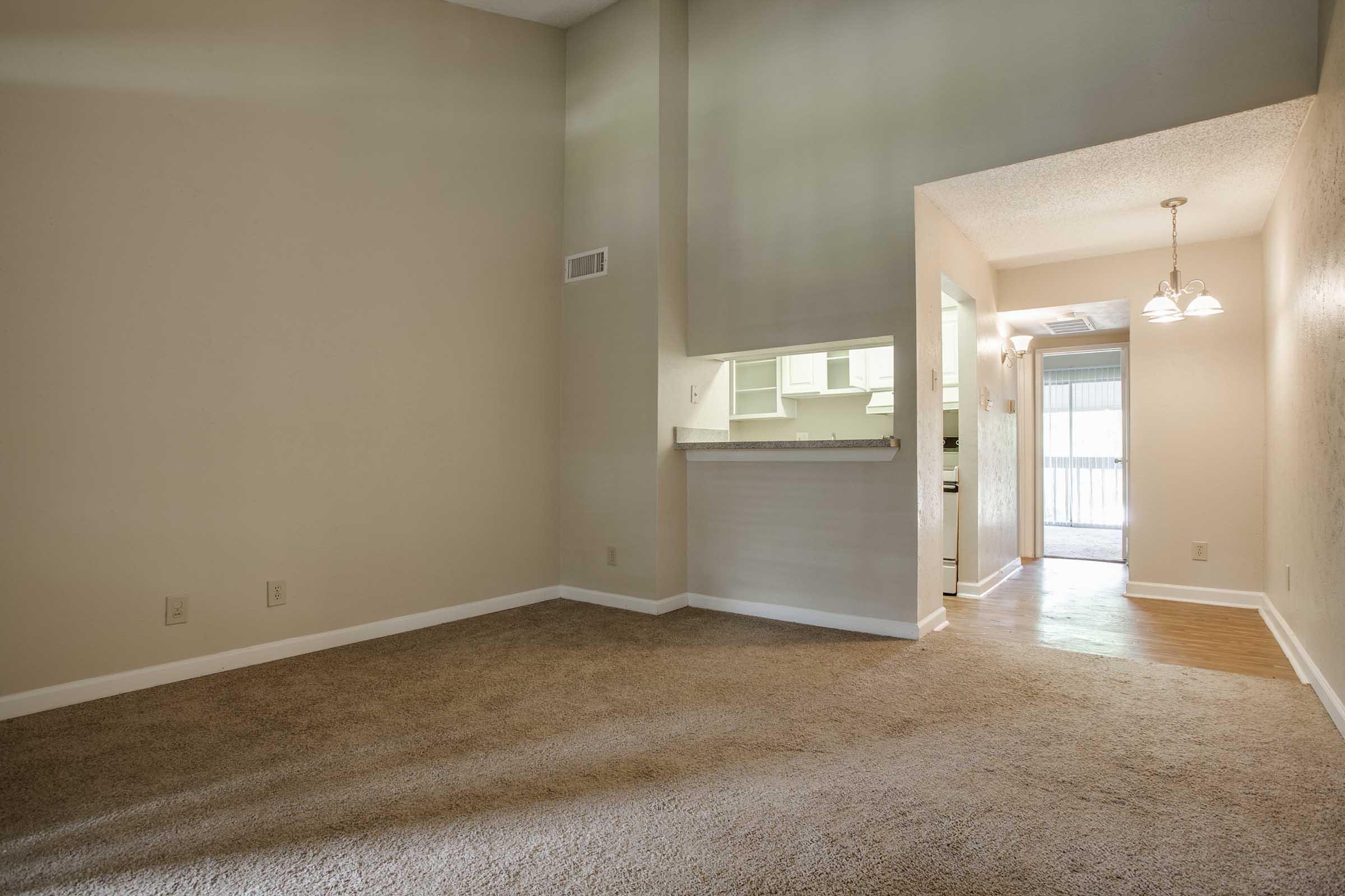 A spacious room with beige carpet and light-colored walls, featuring a small bar area that connects to a kitchen. There is a doorway leading to another room, illuminated by natural light. The space appears open and inviting, with minimal furniture and decor.