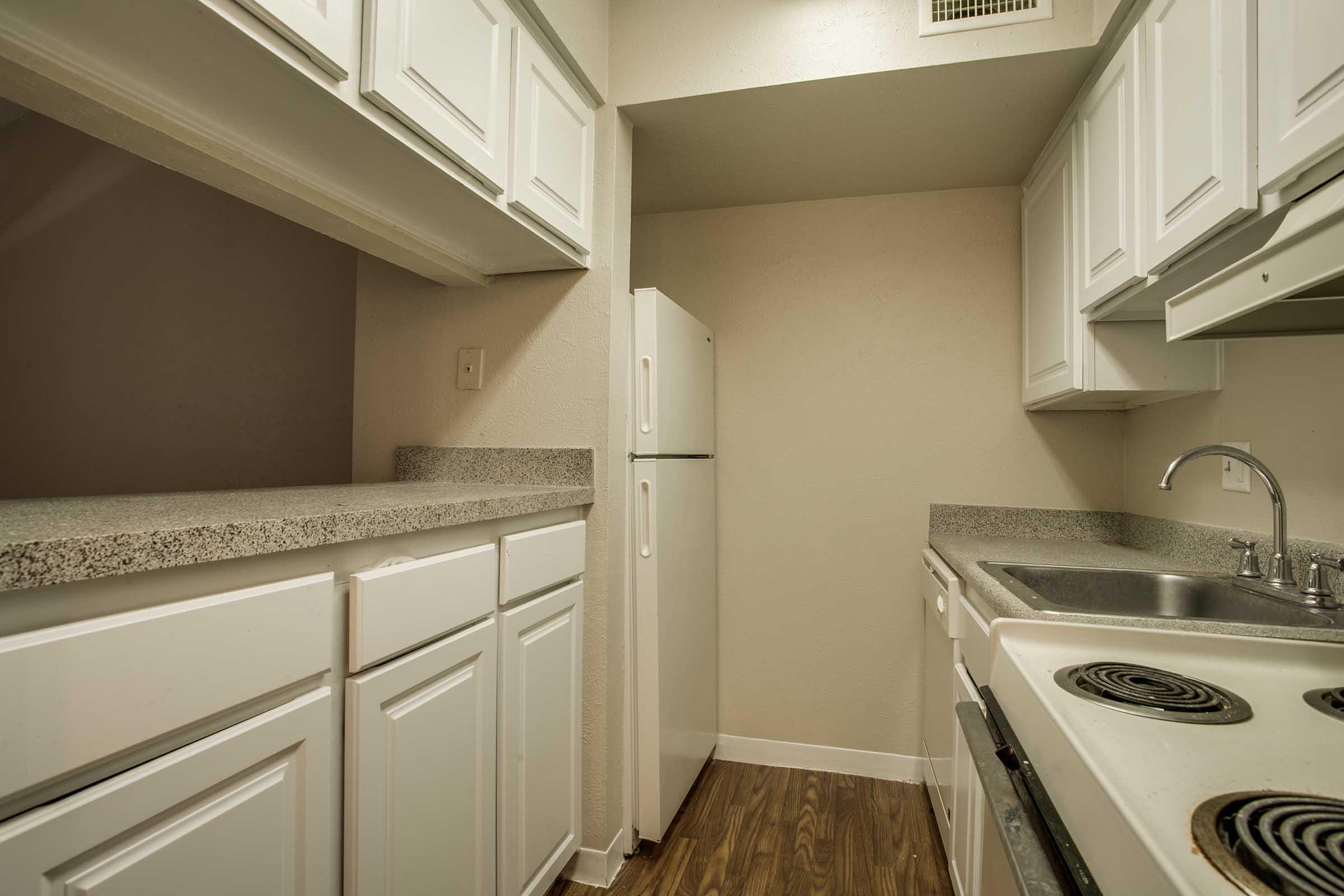 A compact kitchen featuring white cabinetry, a refrigerator, a double basin sink, and a stove with an oven. The countertops are gray, and the flooring is dark wood. The walls are painted in a light color, creating a bright and clean atmosphere.