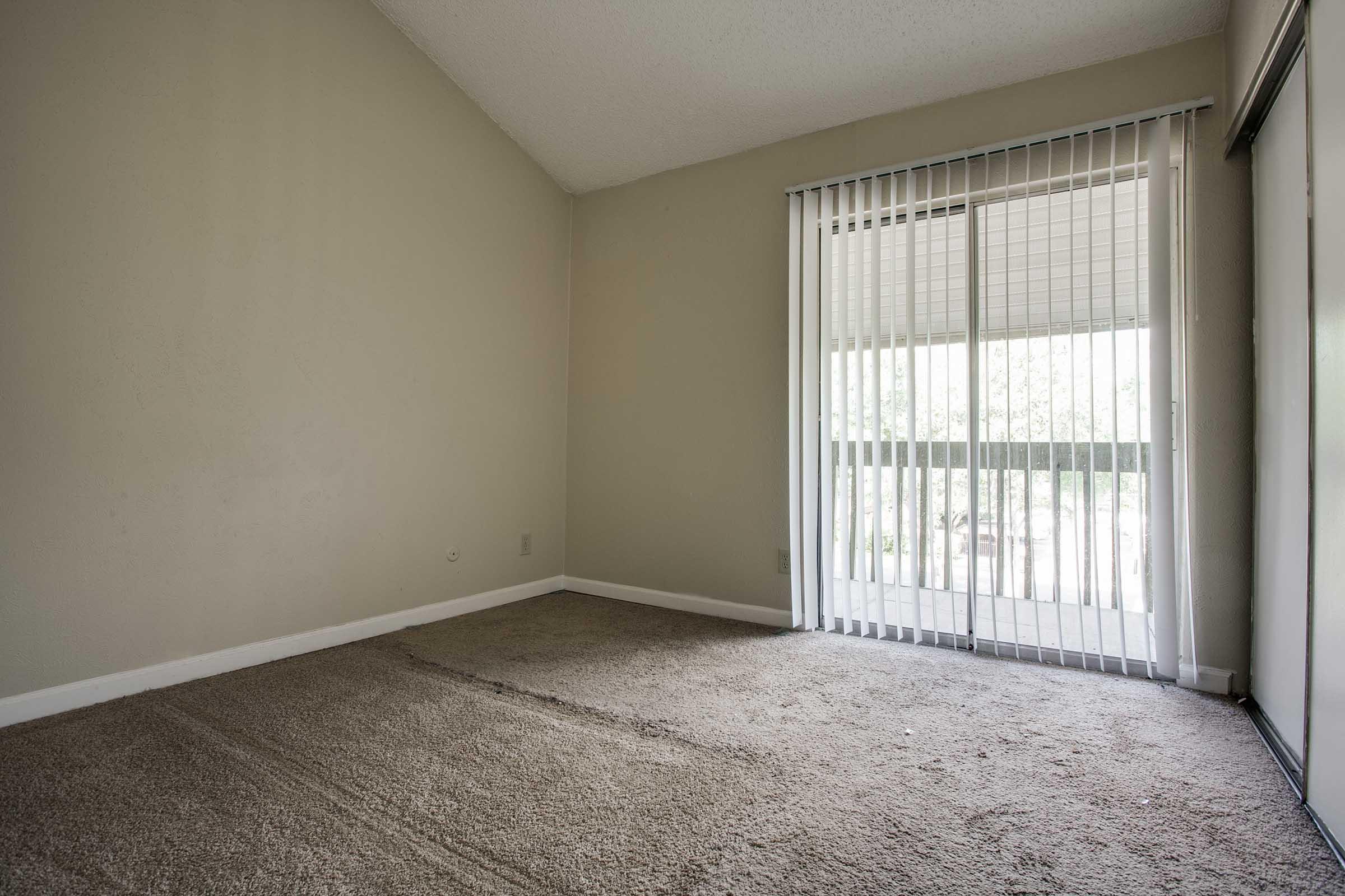Empty room with beige walls and carpeted floor, featuring a sliding glass door with vertical blinds leading to a balcony. The room has a ceiling with a slight angle, creating a spacious feel. Natural light enters through the door, highlighting the plain and uncluttered space.