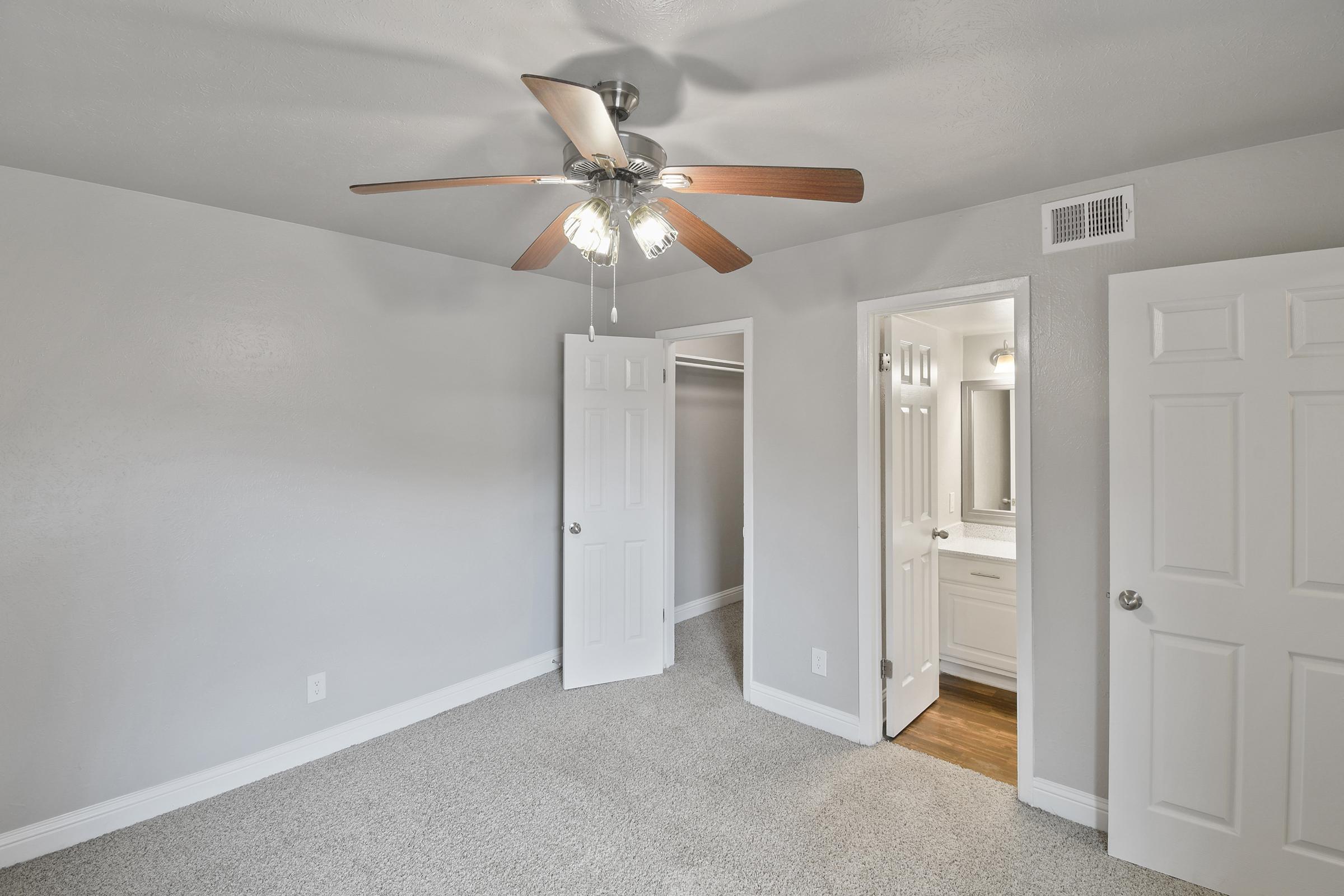 A well-lit, neutral-toned bedroom featuring carpeted flooring, a ceiling fan with wooden blades, and two doors leading to a closet and a bathroom. The walls are painted light gray, and the overall ambiance is spacious and inviting.