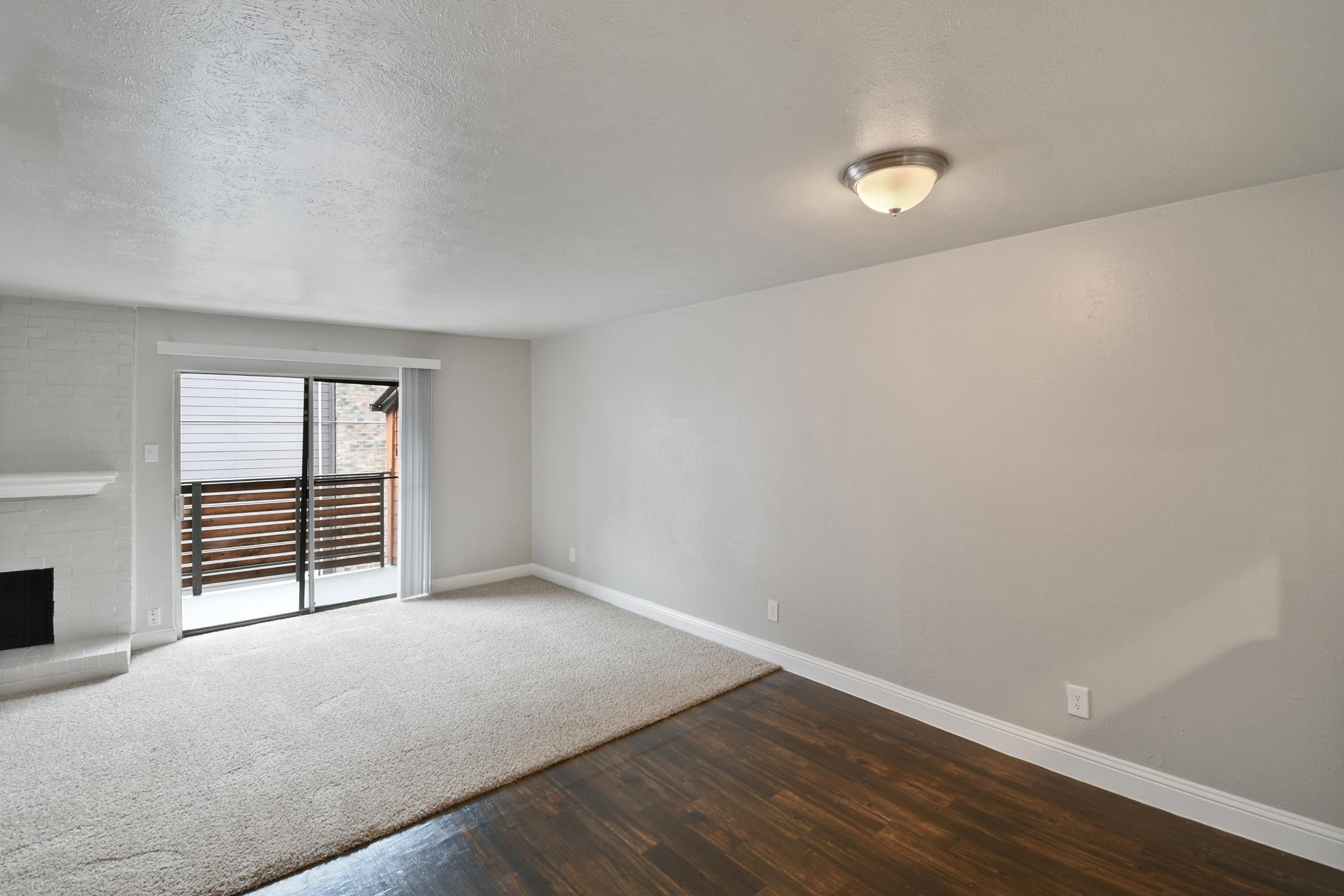 Bright and spacious living room with light beige walls and a combination of carpet and hardwood flooring. A sliding glass door leads to a balcony, allowing natural light to fill the space. A simple ceiling light fixture provides illumination, and there's a fireplace for added warmth.