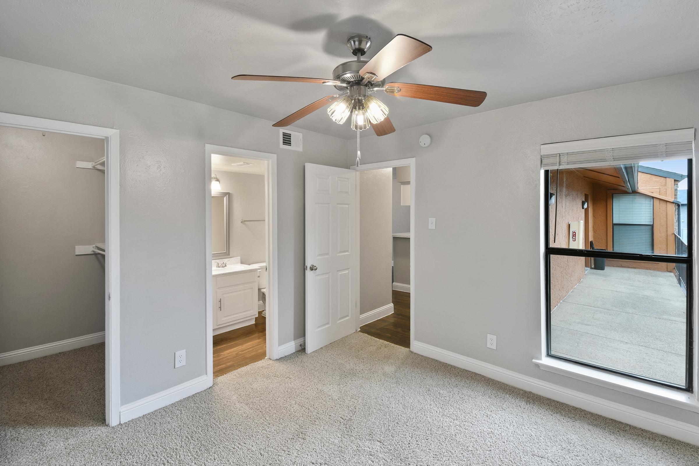 A well-lit bedroom featuring light grey walls and plush carpeting. The room has a ceiling fan with lights, a window with blinds, and two open doorways leading to a bathroom and a closet. Natural light streams in from the window, illuminating the cozy space.