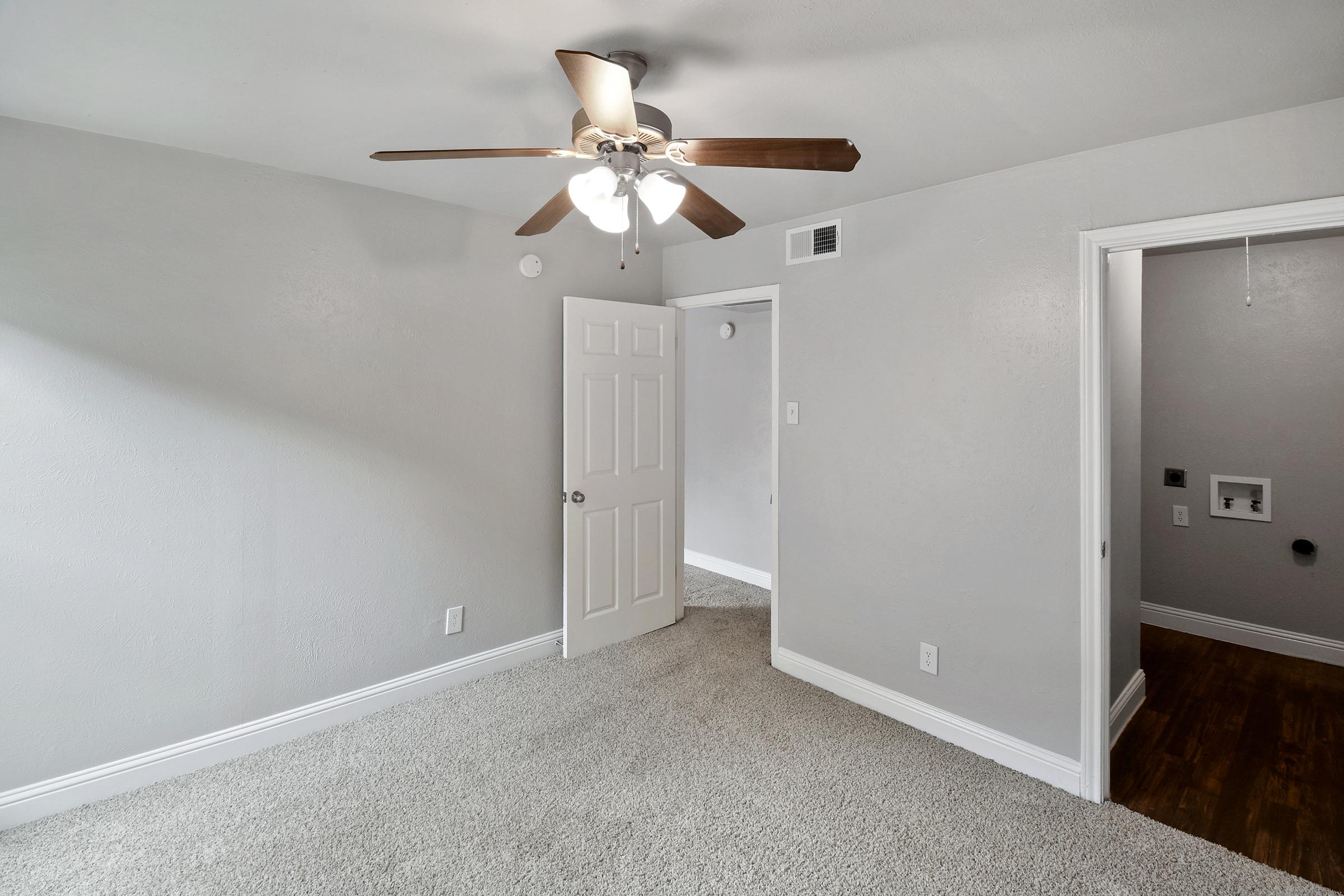 A light gray room featuring a ceiling fan with wooden blades. The floor is covered in beige carpet. There are two doorways: one to the left leading to another space and another straight ahead. The walls are plain and the lighting is bright, creating a clean and spacious feel.