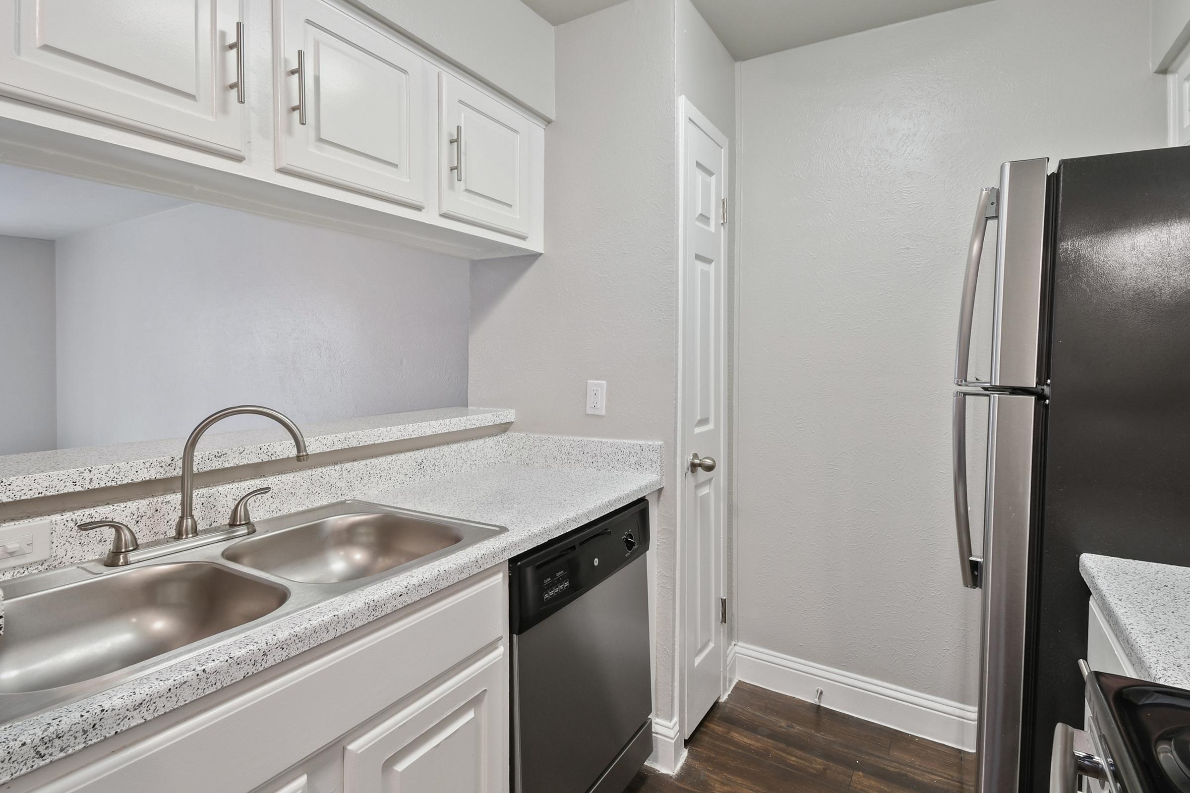 A modern kitchen featuring white cabinetry, a double sink with a brushed steel faucet, and granite countertops. There are stainless steel appliances, including a refrigerator and a dishwasher, set against a neutral gray wall. The flooring is dark wood, creating a contemporary aesthetic.