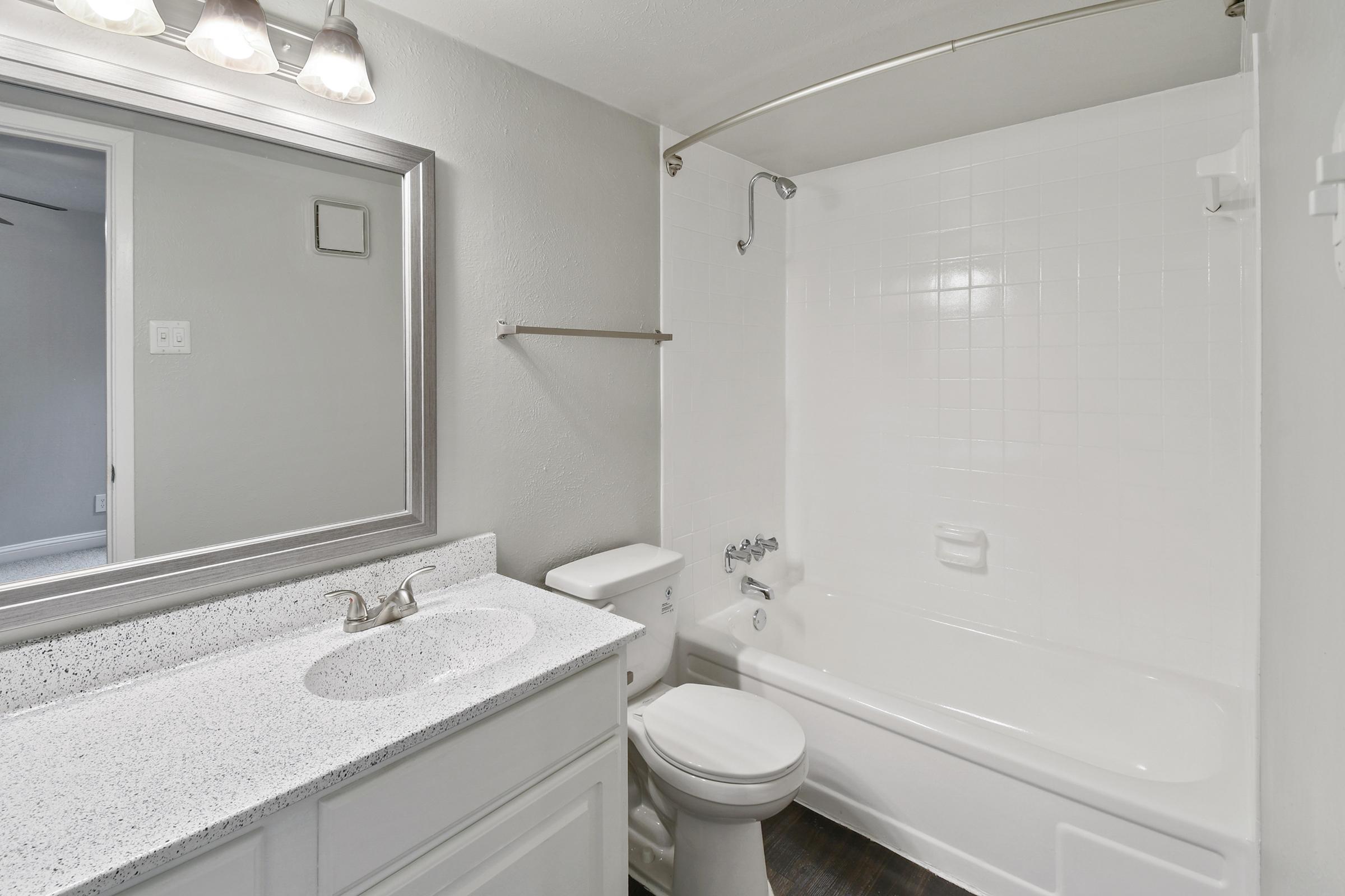 A modern bathroom featuring a white bathtub, a toilet, and a sink with a light-colored countertop. A rectangular mirror is mounted above the sink, and the walls are painted in light grey. The bathroom has a clean and minimalistic design with a neutral color palette.