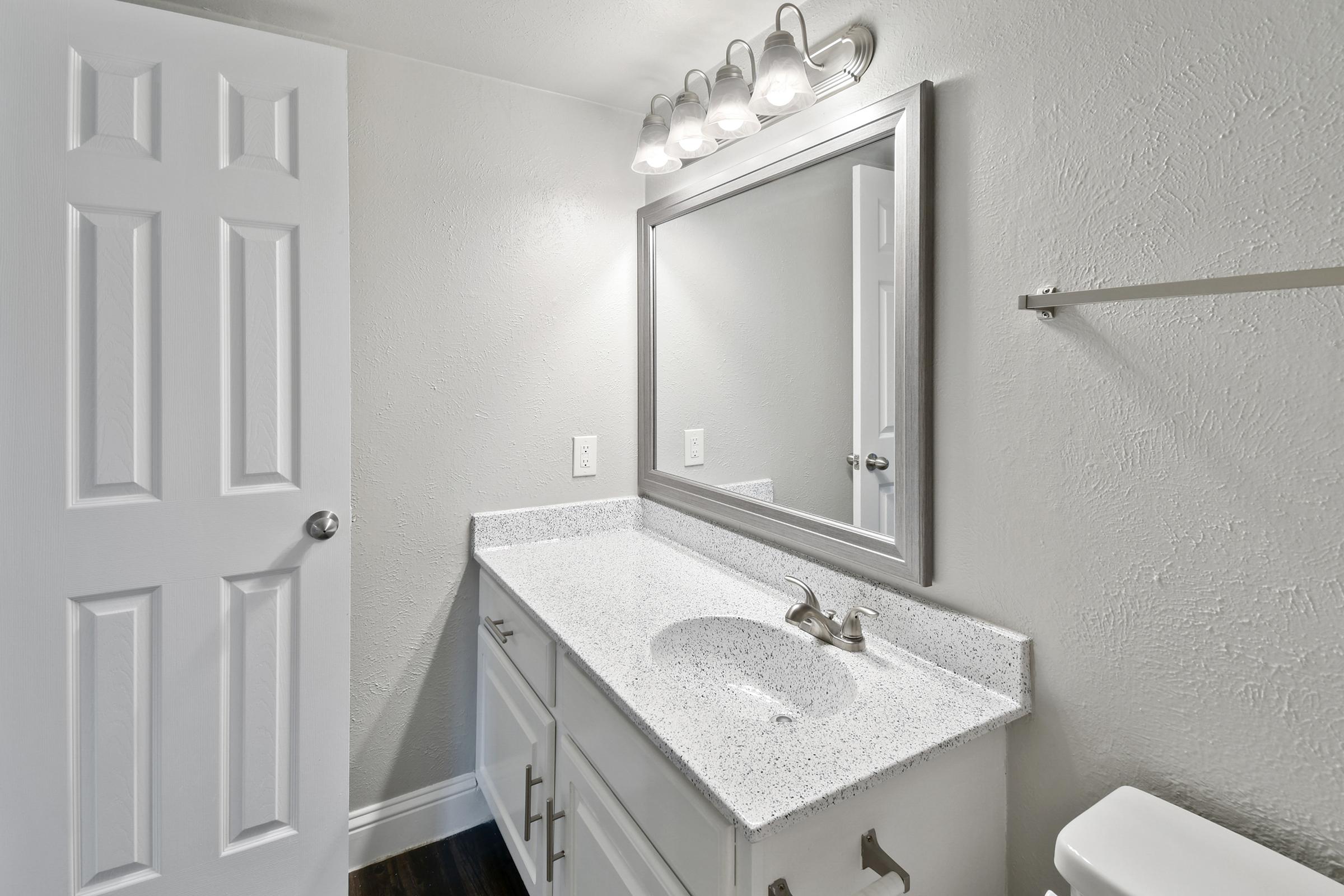 A modern bathroom featuring a white countertop with a sink, a large mirror above it, and a light fixture with multiple bulbs. There is a closed white door on the left, a towel bar on the wall, and a toilet visible in the corner, all set against light gray walls and dark flooring.