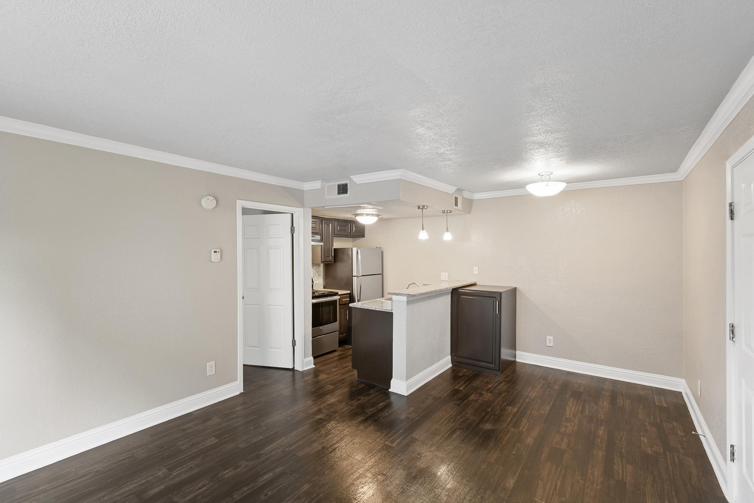 A spacious living area with hardwood flooring, featuring a kitchenette with dark cabinets and modern appliances. The walls are painted in a neutral tone, and there are pendant lights above the kitchen island. A door leads to the hallway, and white crown molding adds a decorative touch.