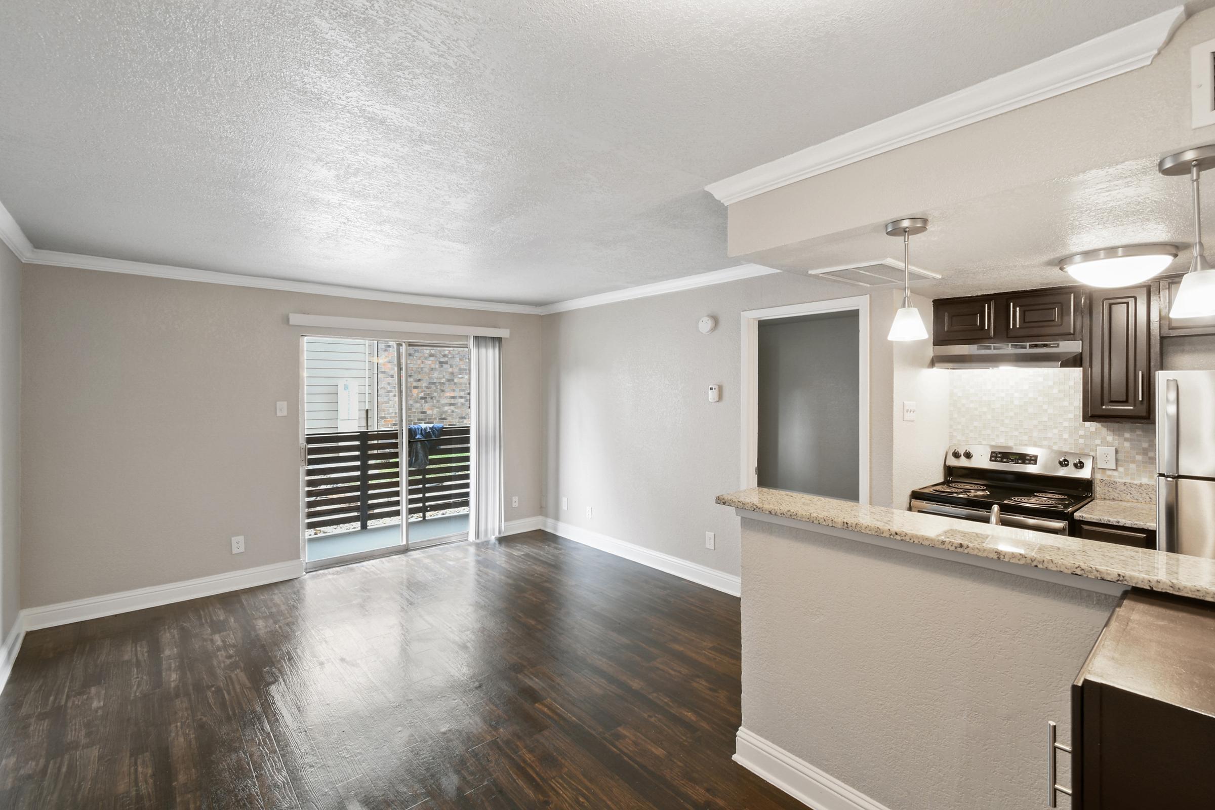 Interior view of a modern apartment featuring an open floor plan with dark wooden flooring, light-colored walls, and a kitchen area with dark cabinets. There are sliding glass doors leading to a balcony, and the kitchen includes stainless steel appliances and a breakfast bar. Natural light fills the space.