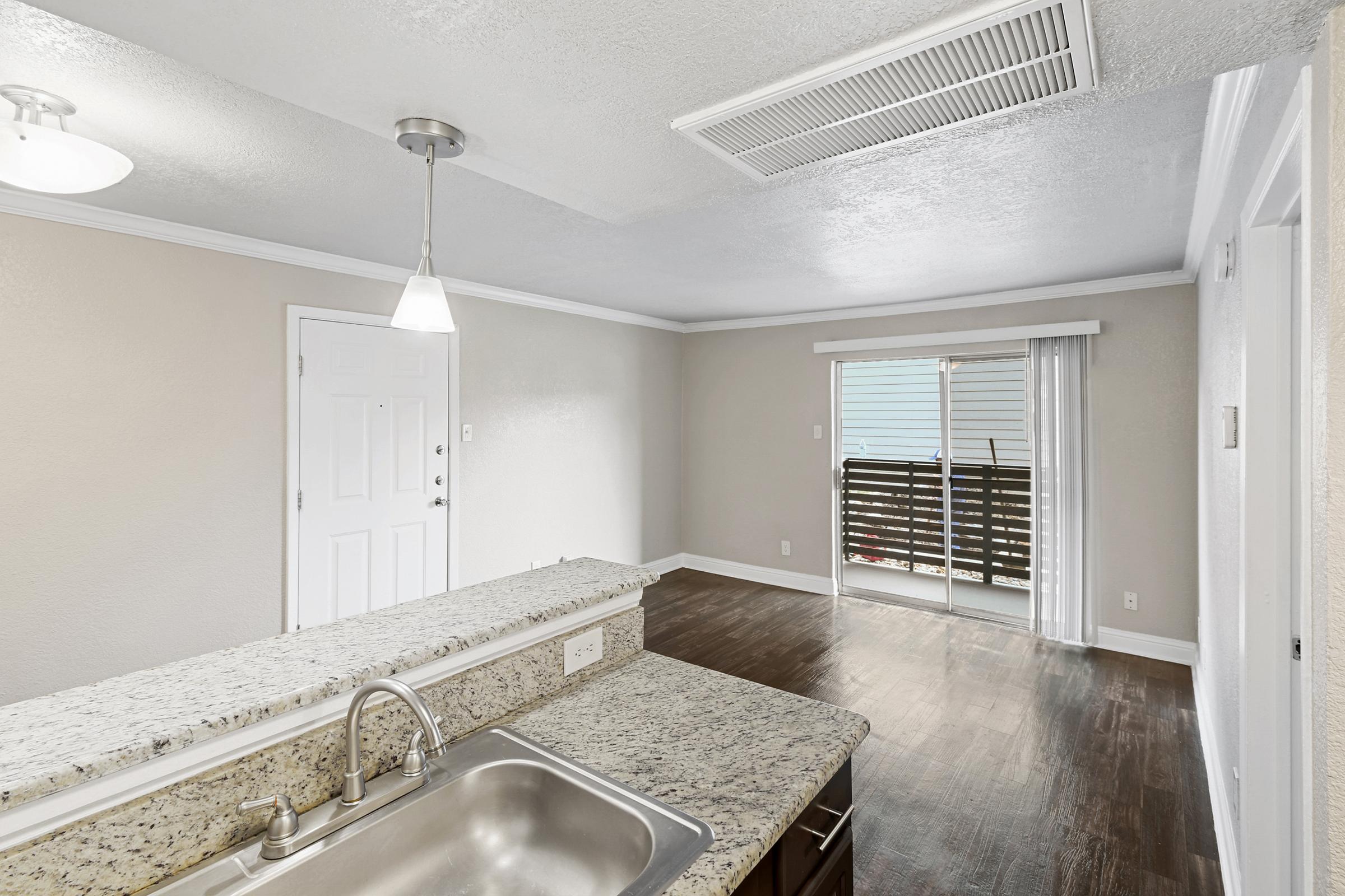Interior view of a modern living space featuring a granite kitchen countertop, a sink with a faucet, and an open layout leading to a balcony. Natural light enters through sliding glass doors, and there is a ceiling light fixture and air vent visible. The walls have a light neutral color.