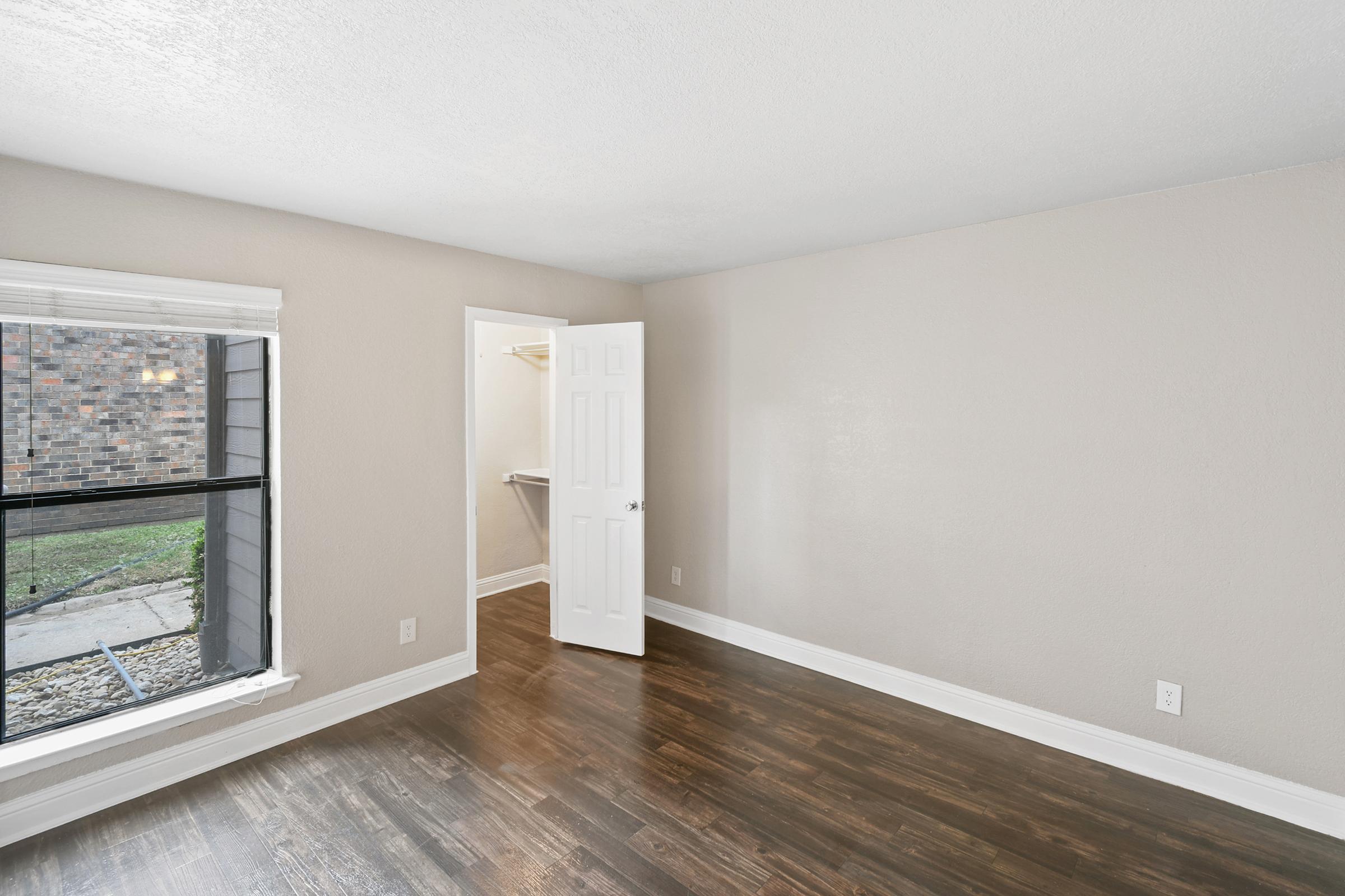 A bright, empty room featuring light gray walls and dark wooden flooring. There is a large window on one wall, allowing natural light to enter. An open door leads to a closet area, and the overall space appears clean and well-maintained, suitable for various uses.