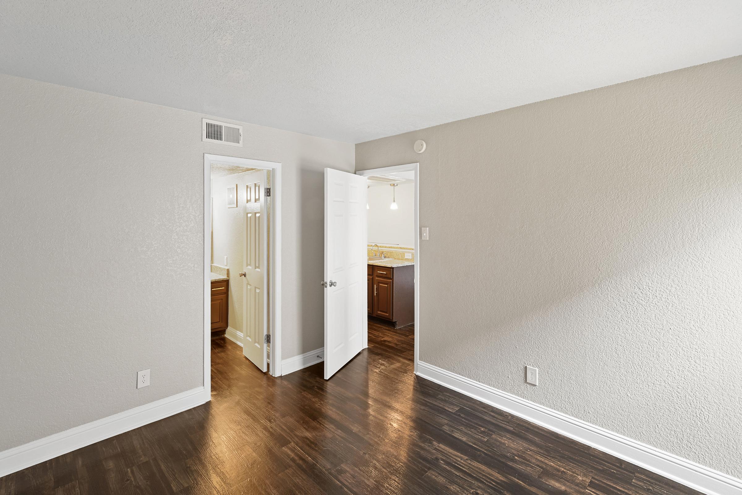 A well-lit room featuring light gray walls and dark wood flooring. A partially opened door leads to a bathroom area with brown cabinetry. The space appears fresh and modern, with a clean, uncluttered look. Natural light enters from an unseen source, enhancing the inviting atmosphere.
