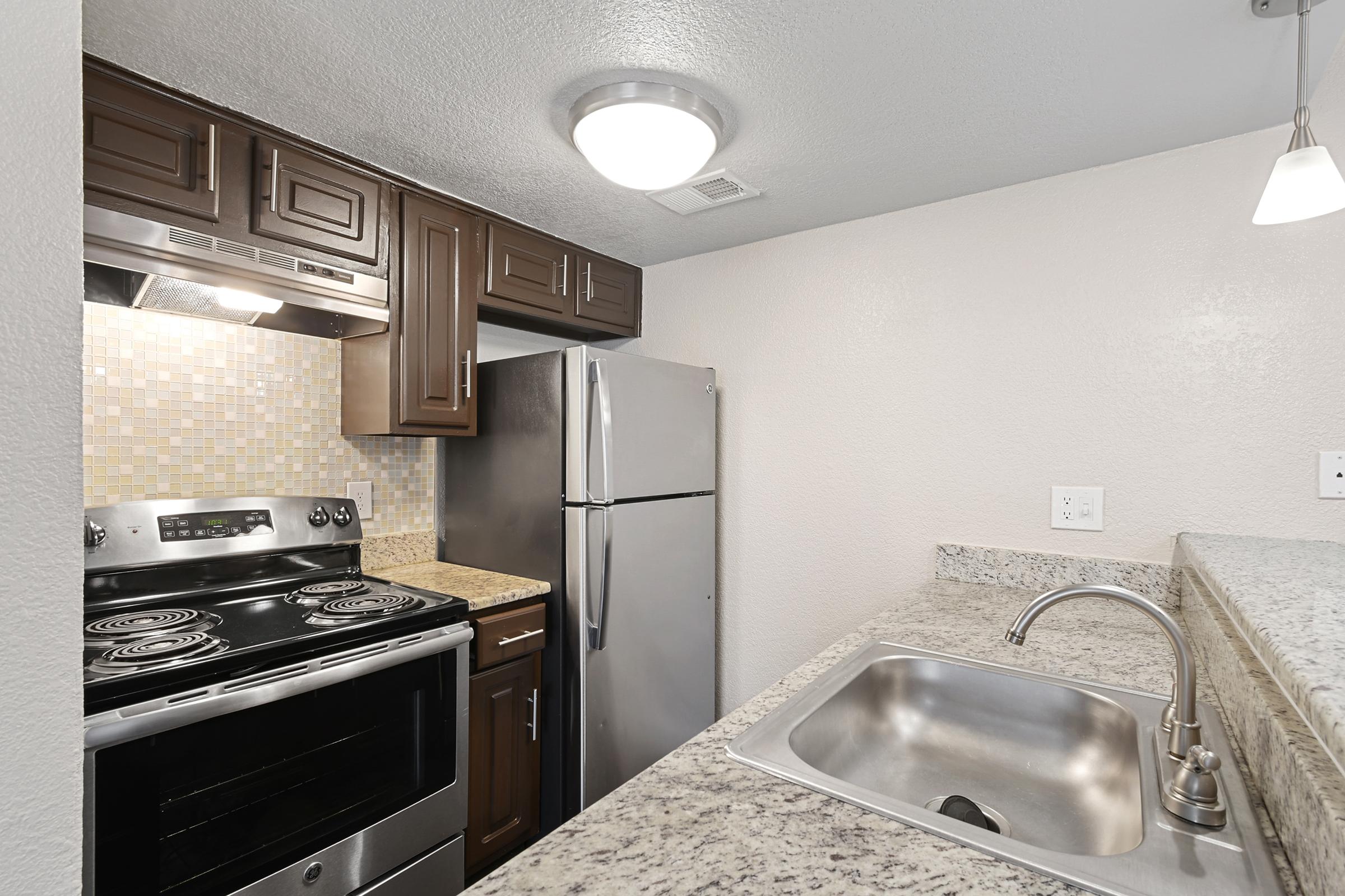 Modern kitchen featuring dark wooden cabinets, a stainless steel refrigerator, an oven with a stove, and a granite countertop. The backsplash is made of textured tiles, and there is a stainless steel sink with a sleek faucet. Soft lighting enhances the contemporary feel of the space.
