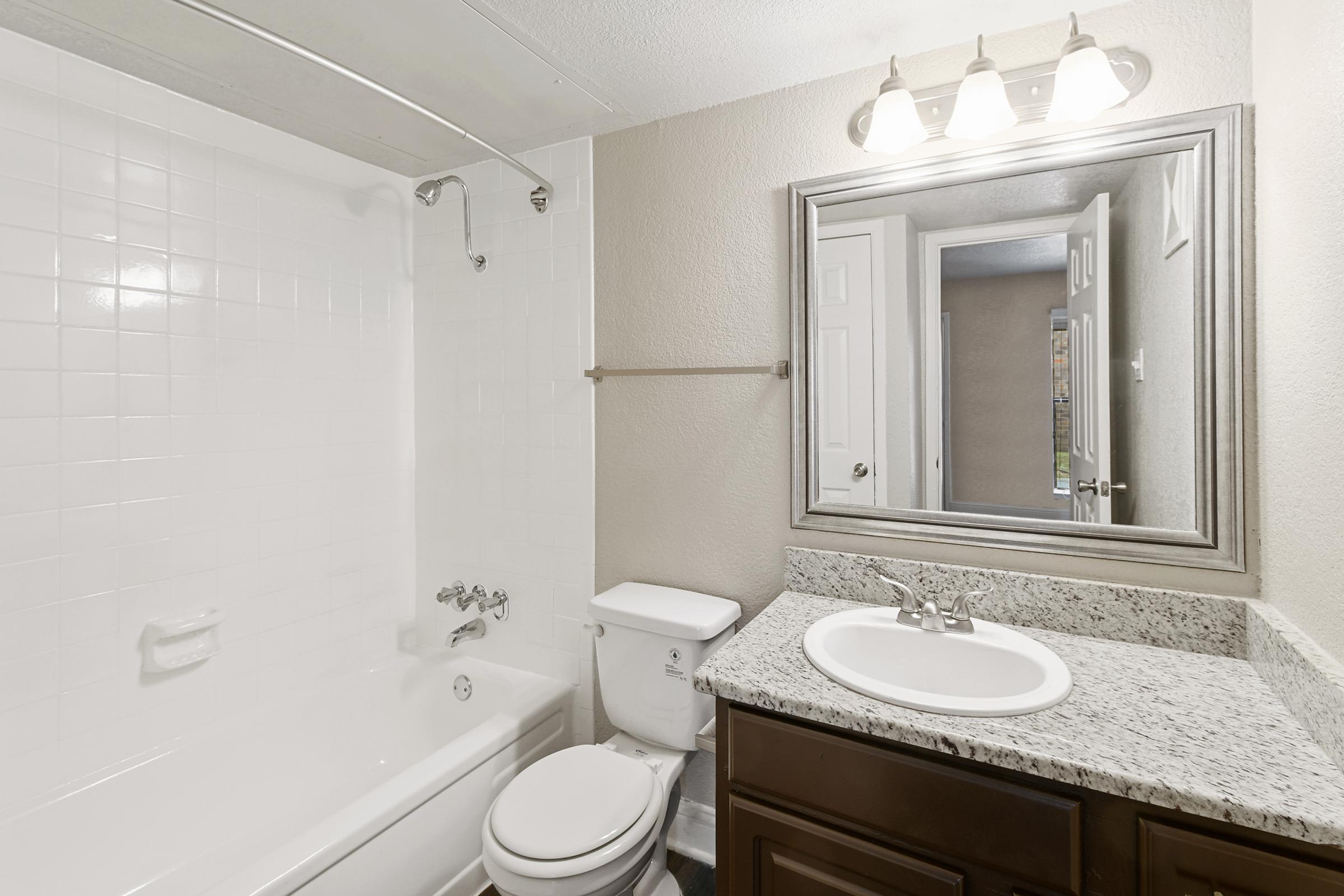 A clean and well-lit bathroom featuring a white tiled shower/tub combination, a modern vanity with a granite countertop, a round sink, and a mirror with three light fixtures above. The walls are painted in a neutral color, and a door leads to another room.