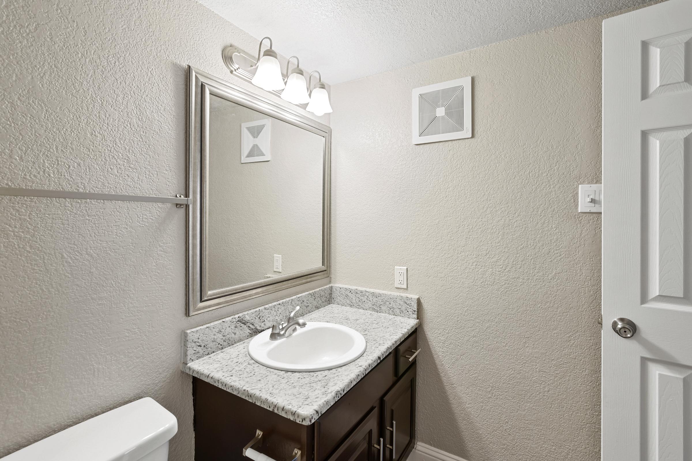 A well-lit bathroom featuring a white countertop with a sink, a large mirror above it, and a gray textured wall. There is a light fixture with three bulbs above the mirror, and a door on the right side. A simple framed artwork is hung above the sink, adding a touch of decor to the space.