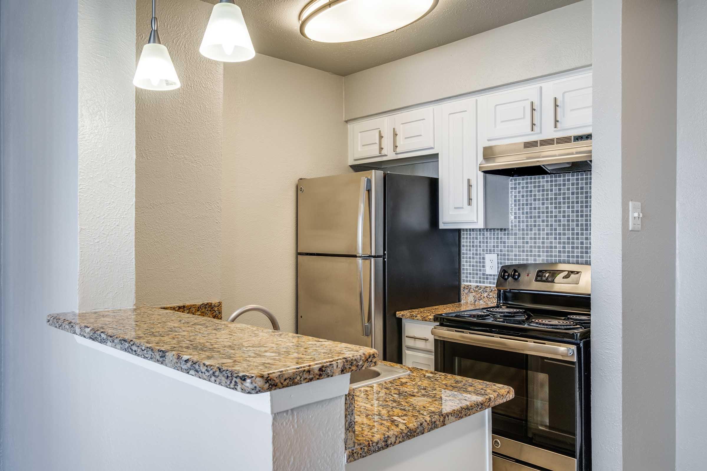 Modern kitchen space featuring white cabinets, stainless steel appliances including a refrigerator and oven, granite countertops, and a tiled backsplash. There are two pendant lights above the counter area, enhancing the bright and functional design.