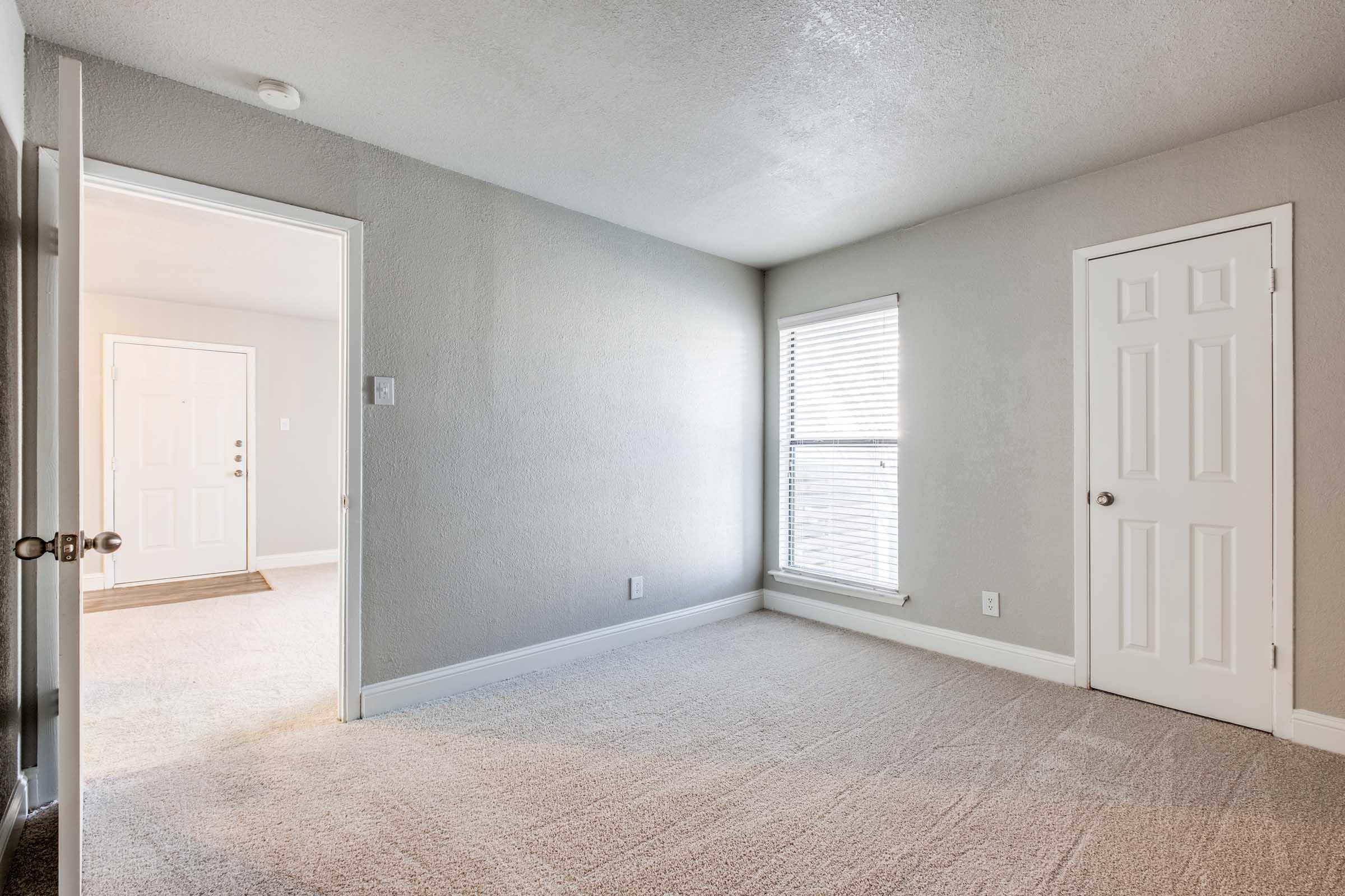 A well-lit, empty room with neutral gray walls and plush carpeting. One window lets in natural light, and there are two doors: one leading to a hallway and the other closed. The room appears clean and spacious, suitable for various furniture arrangements.