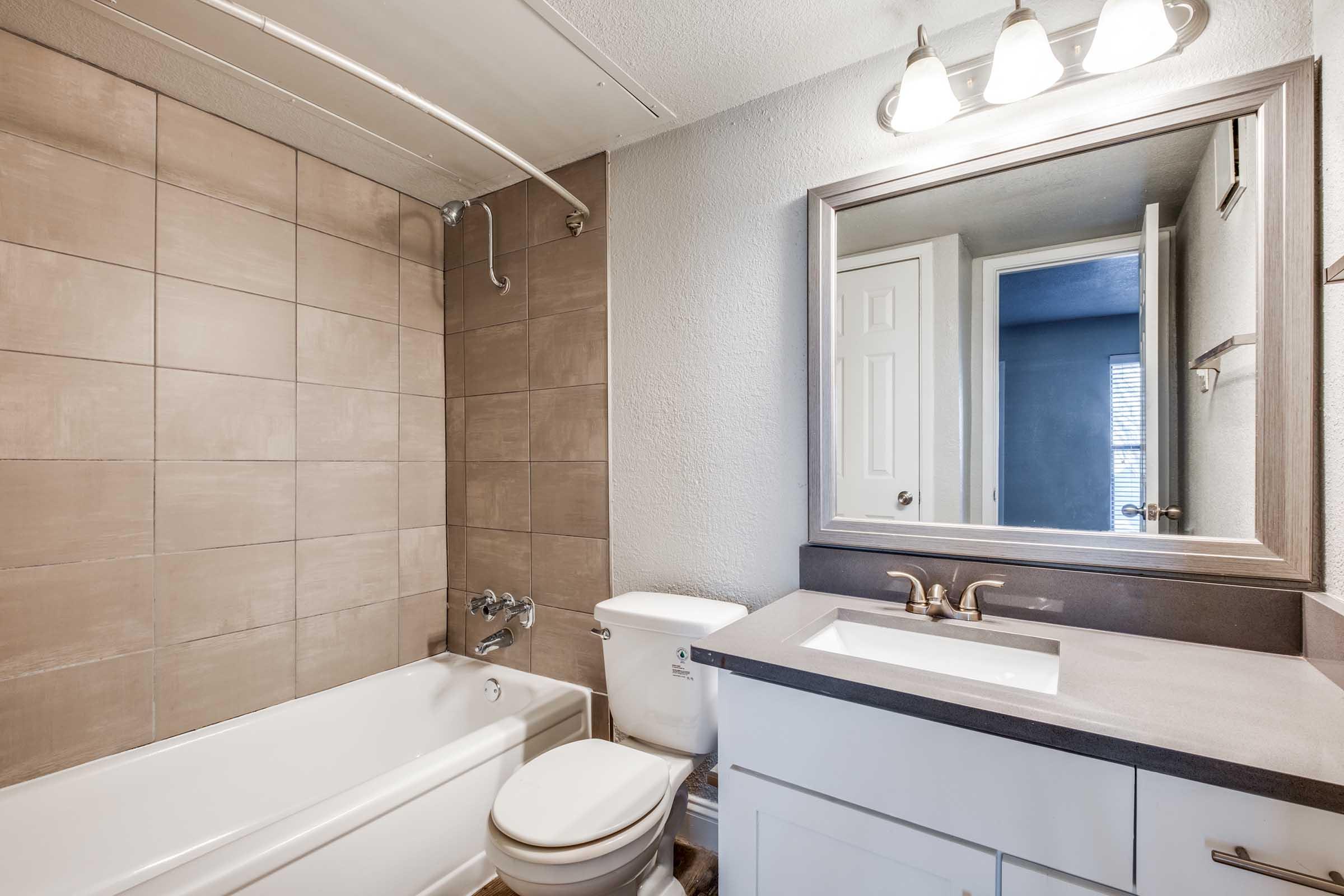 A modern bathroom featuring a bathtub with a shower, tiled walls in neutral tones, a white toilet, and a vanity with a sink. A large mirror is mounted above the sink, and there is a door leading to another room in the background. Bright lighting enhances the space.