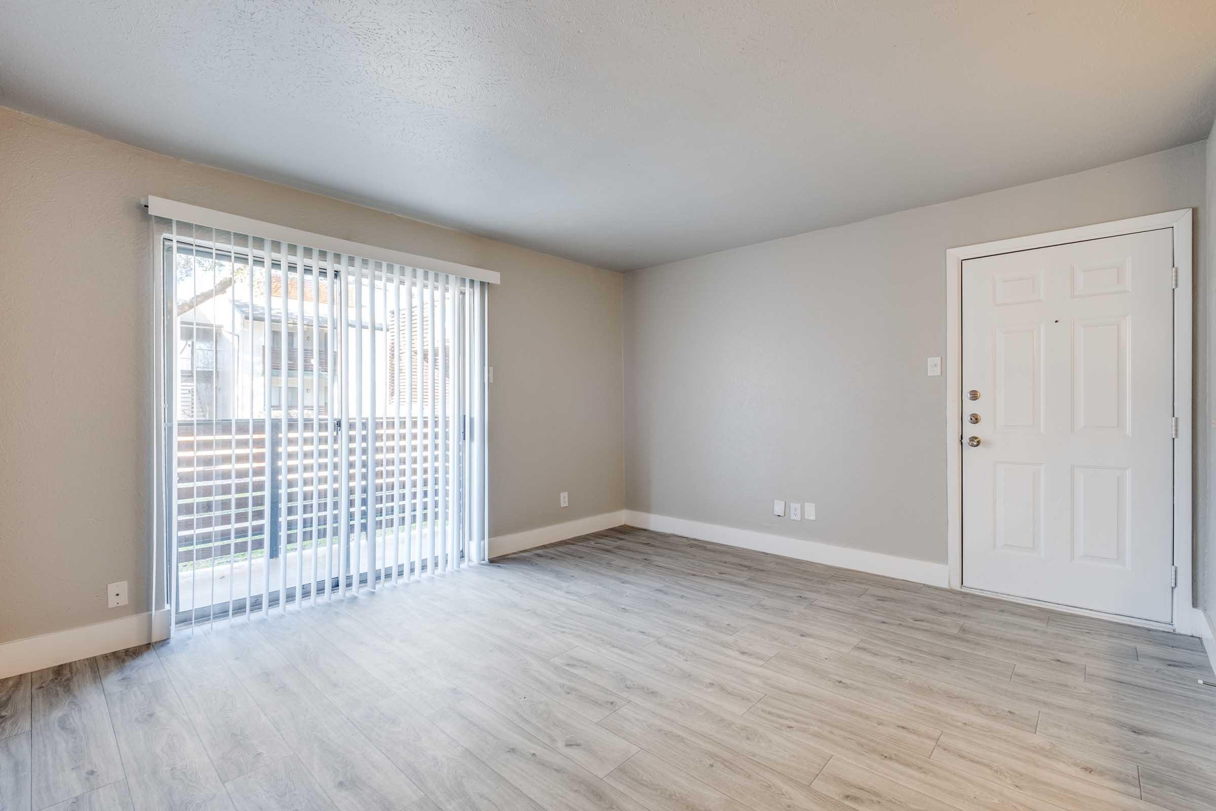 A spacious, empty living room featuring light gray walls and new laminate flooring. There is a sliding glass door with vertical blinds leading to a balcony. A white door with a doorknob is visible on the right side of the room. Natural light fills the area, creating a bright and open atmosphere.