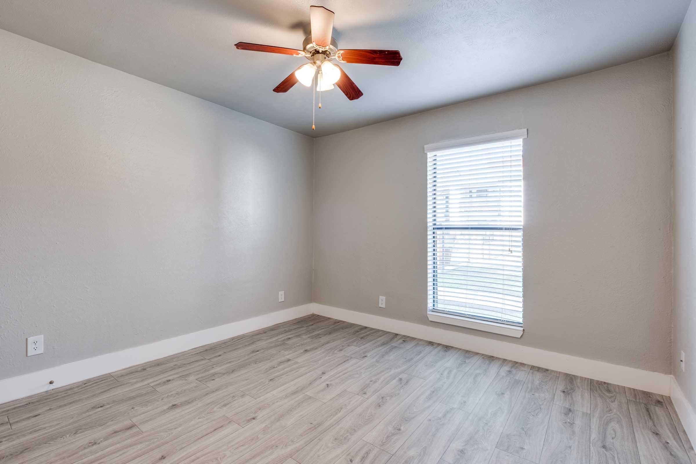 A vacant room with light gray walls, a ceiling fan with wooden blades, and a window dressed with white blinds. The floor is covered in light wood-colored laminate. The room appears bright and spacious, suitable for various uses.