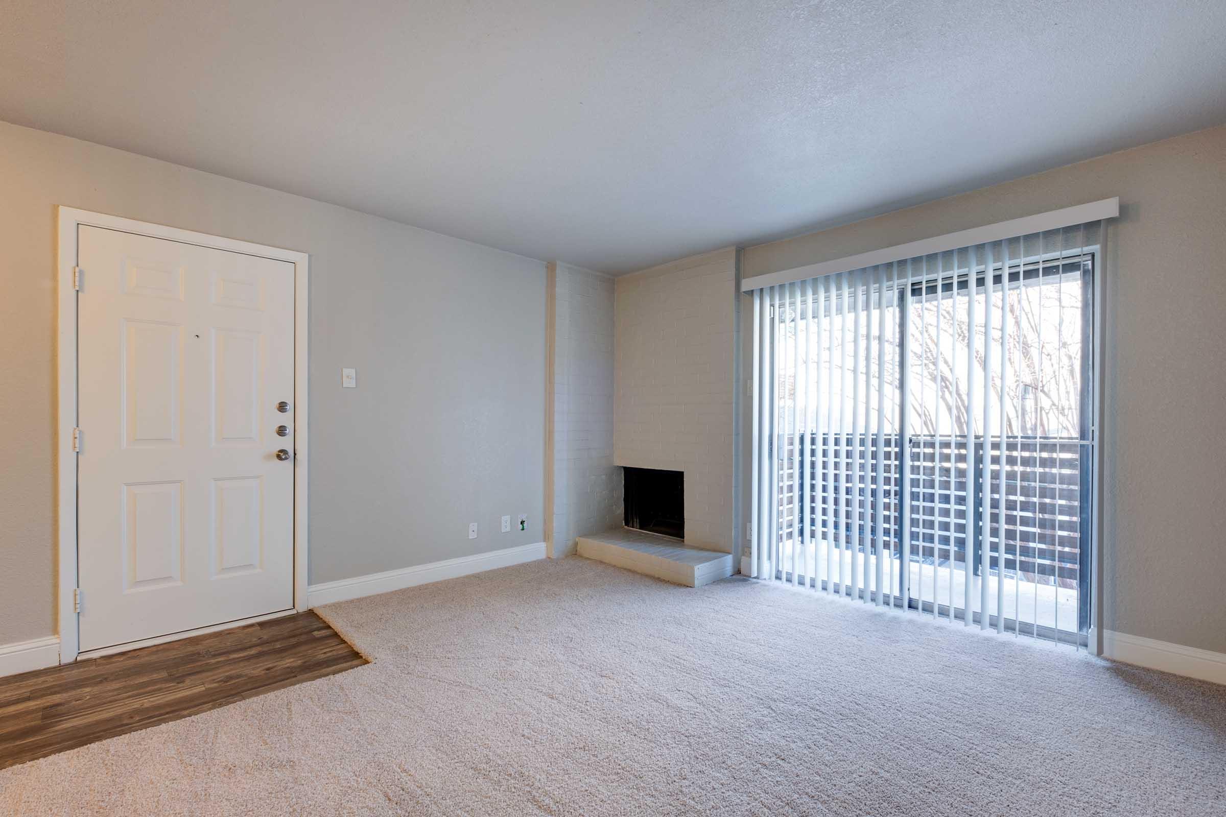 A spacious, empty living room with light beige carpet and gray walls. To the left, a white front door with a doorknob and multiple locks. On the right, a sliding glass door leads to a balcony, with vertical blinds covering the window. A small fireplace is located on the wall opposite the door.