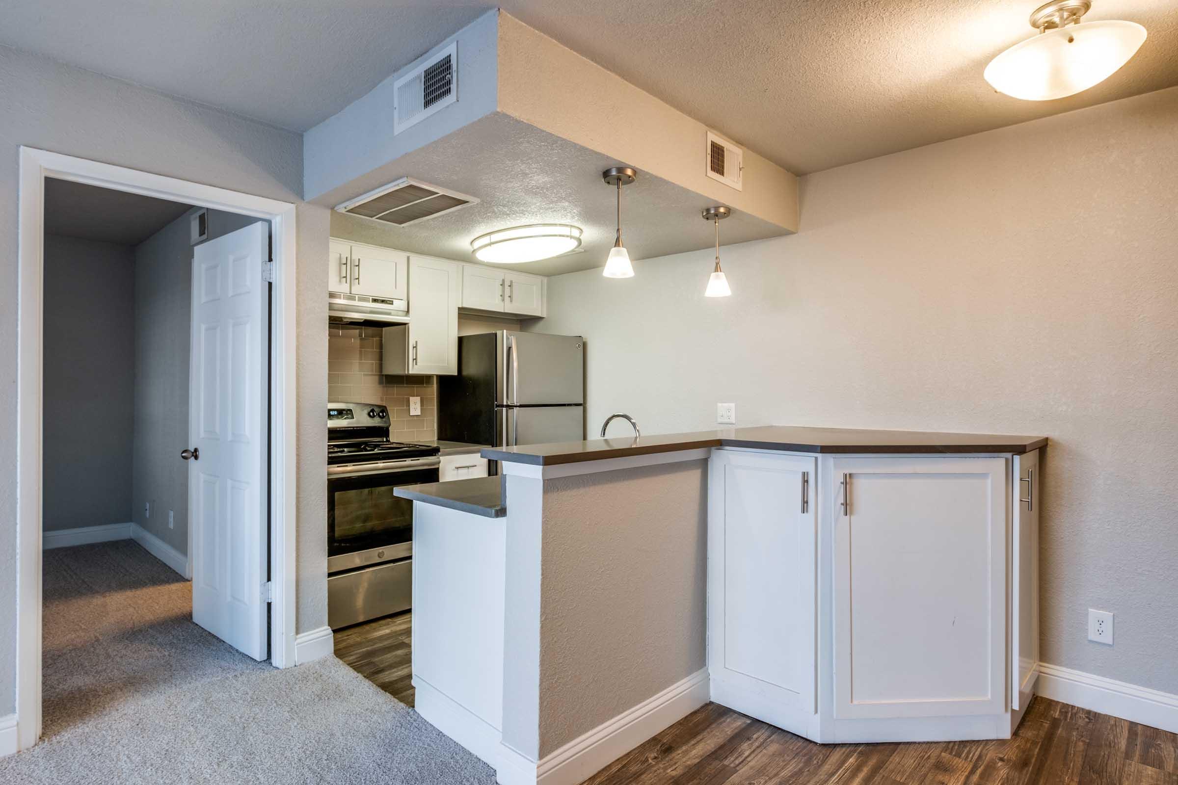 Modern kitchen area showcasing stainless steel appliances, a curved breakfast bar, and pendant lighting. The room features light gray walls and wood-like flooring, with a door leading to an adjacent space. Brightly lit with a ceiling fixture, creating a welcoming atmosphere.