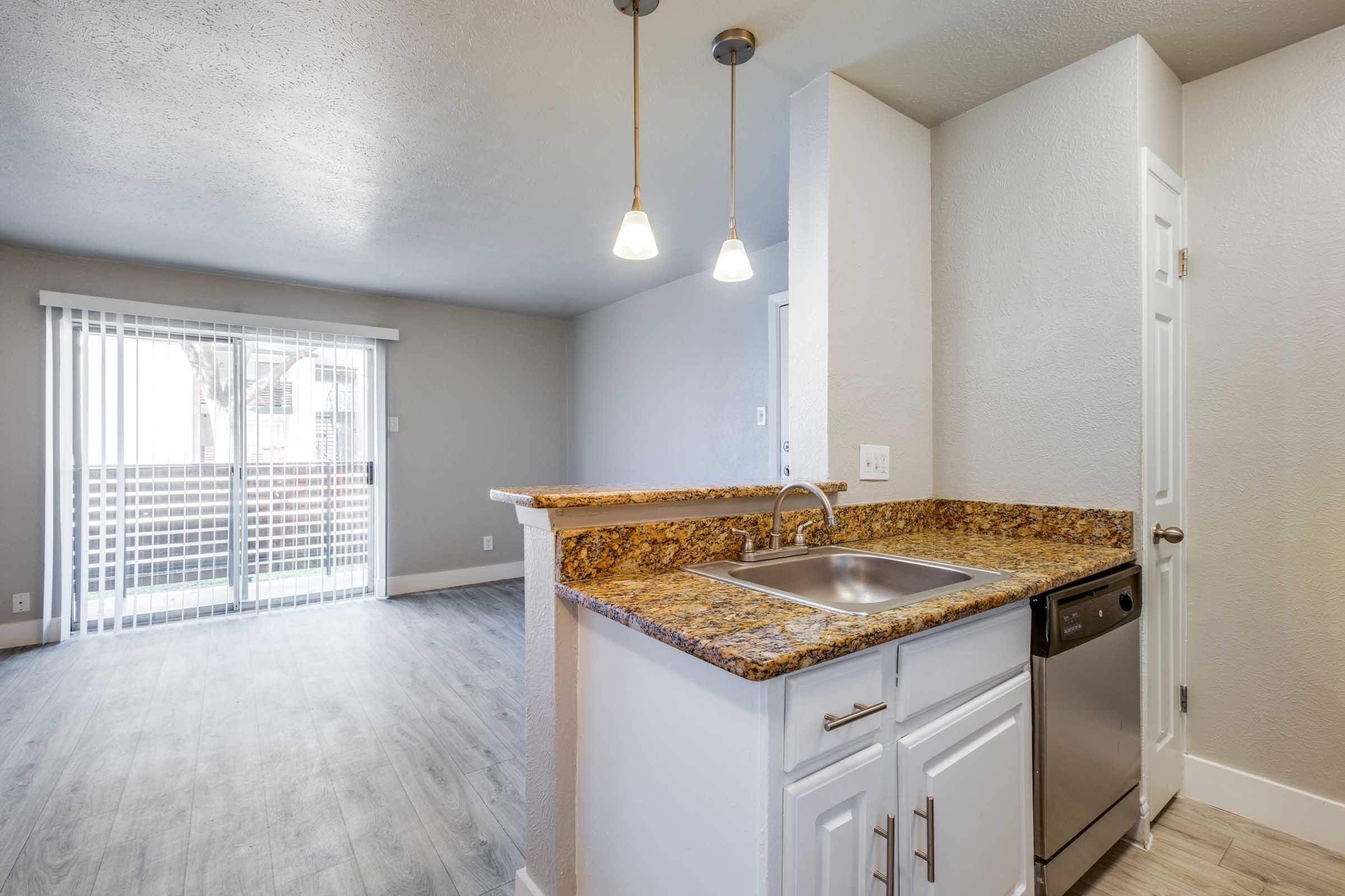 A modern kitchen area with a granite countertop, sink, and dishwasher. The space features pendant lighting and an open layout leading to a living area with sliding glass doors that open to a balcony. The floor is light-colored, enhancing the bright and airy feel of the room.