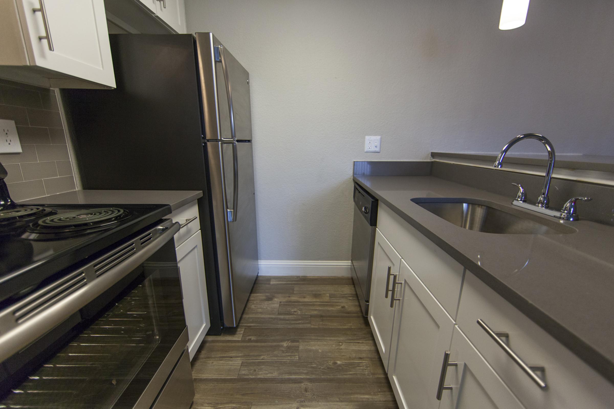 A modern kitchen featuring stainless steel appliances, including a refrigerator and oven. The countertops are a sleek gray, and the cabinets are white. There is a sink with a faucet next to the dishwasher. The floor is wood-like, adding warmth to the contemporary design.