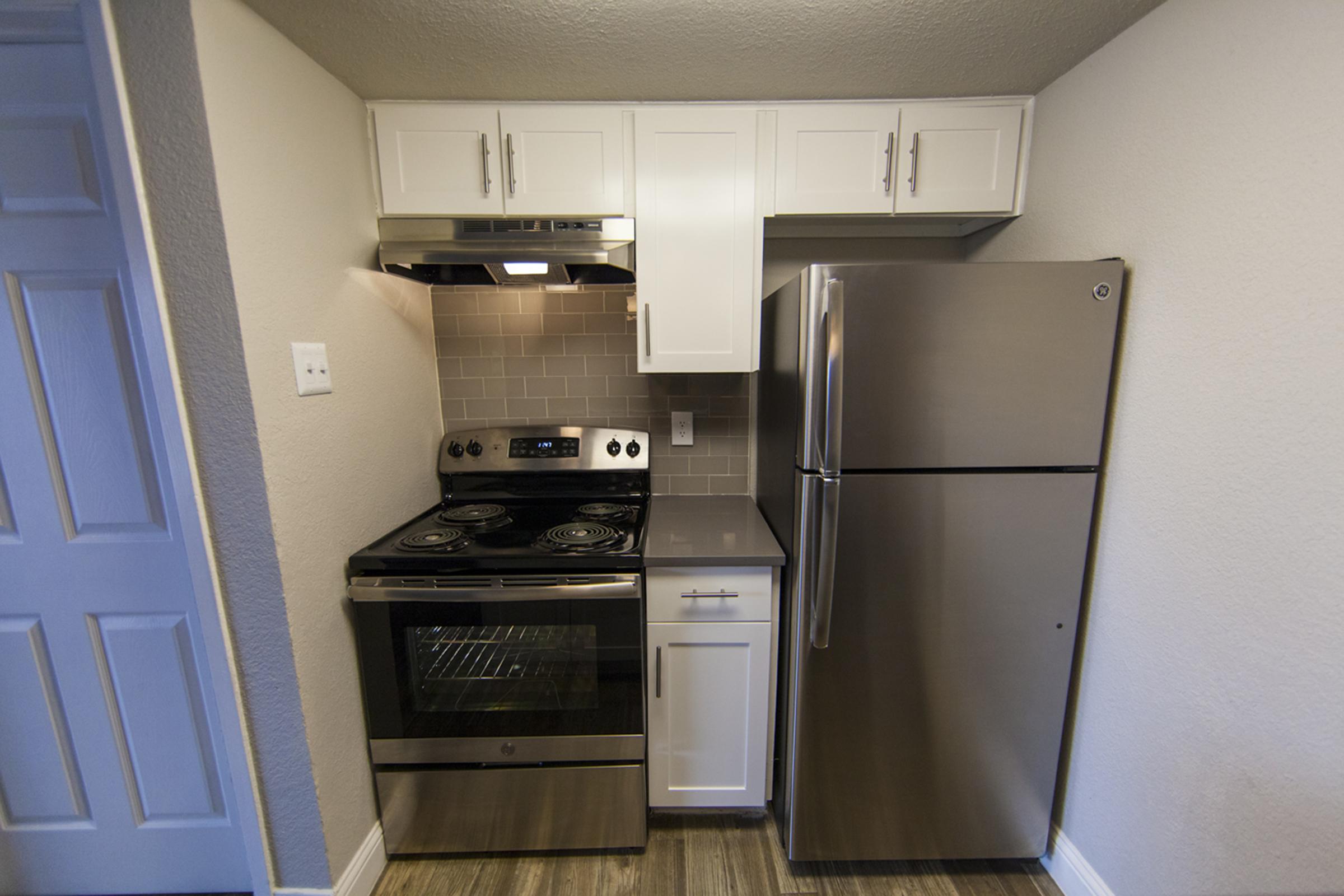 A modern kitchen featuring a stainless steel refrigerator, a black and stainless steel oven with a four-burner cooktop, and white cabinetry. The walls have a light gray color, and there's a tiled backsplash behind the stove. The flooring is wood-like and adds warmth to the space.