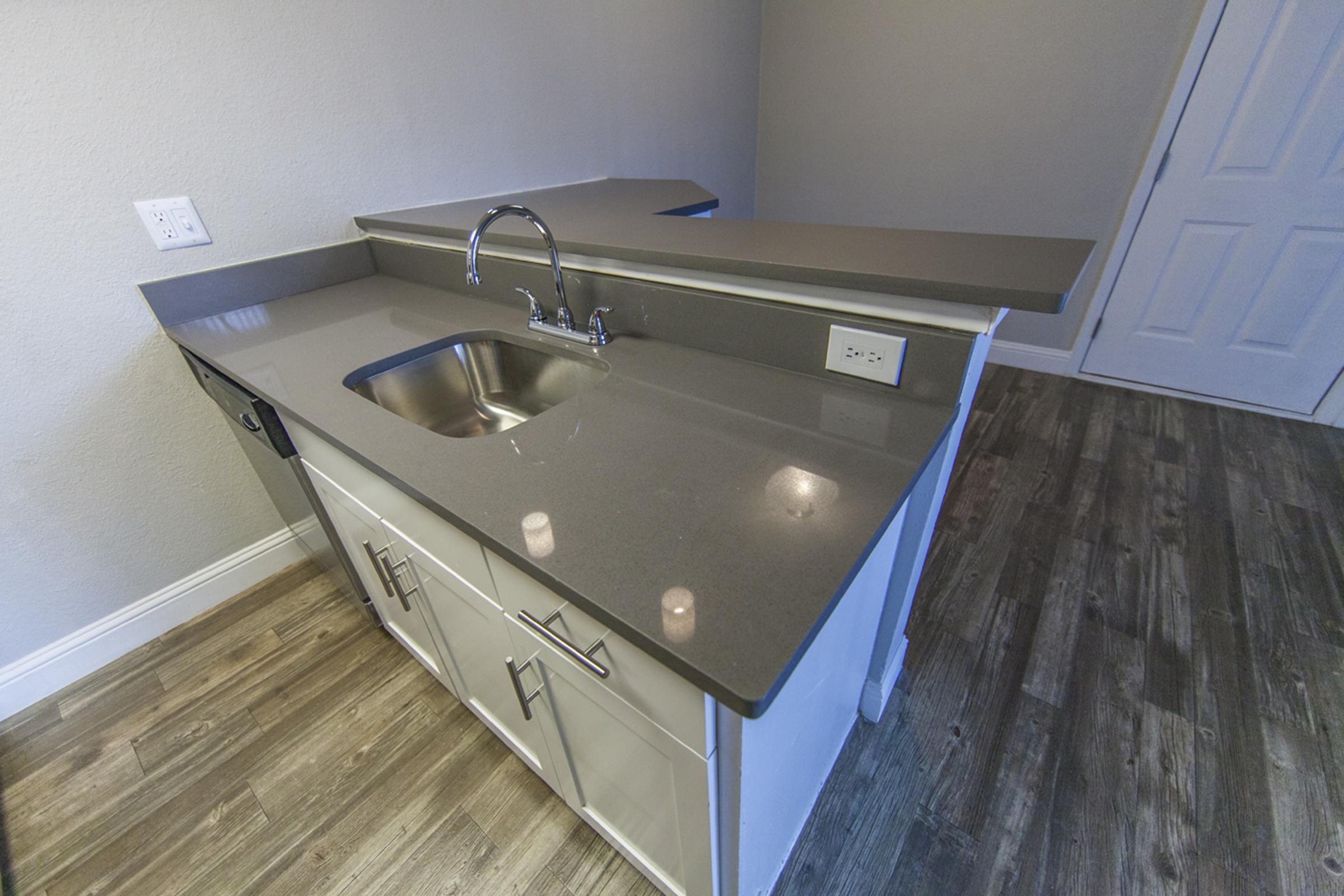 A modern kitchen countertop with a stainless steel sink, featuring sleek gray surfaces. The cabinetry below is designed in a minimalist style with white doors. The floor showcases wood-like laminate, enhancing the contemporary feel, while the walls are painted in a light color. A doorway is visible in the background.