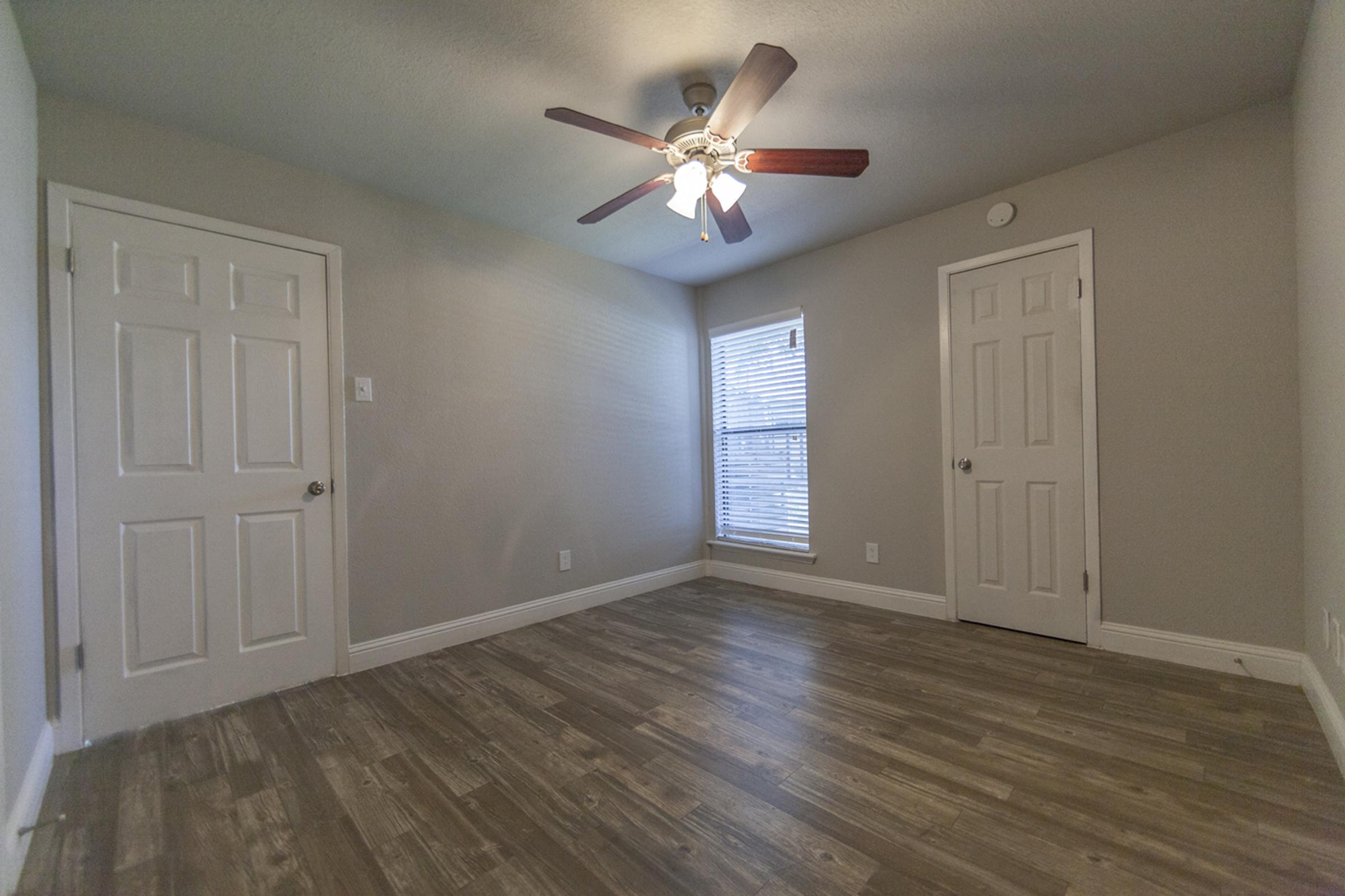 Empty room with light gray walls and wooden flooring. A ceiling fan with wooden blades hangs from the ceiling. Two closed white doors are visible on the left and right side of the image. A window with blinds allows natural light into the space.