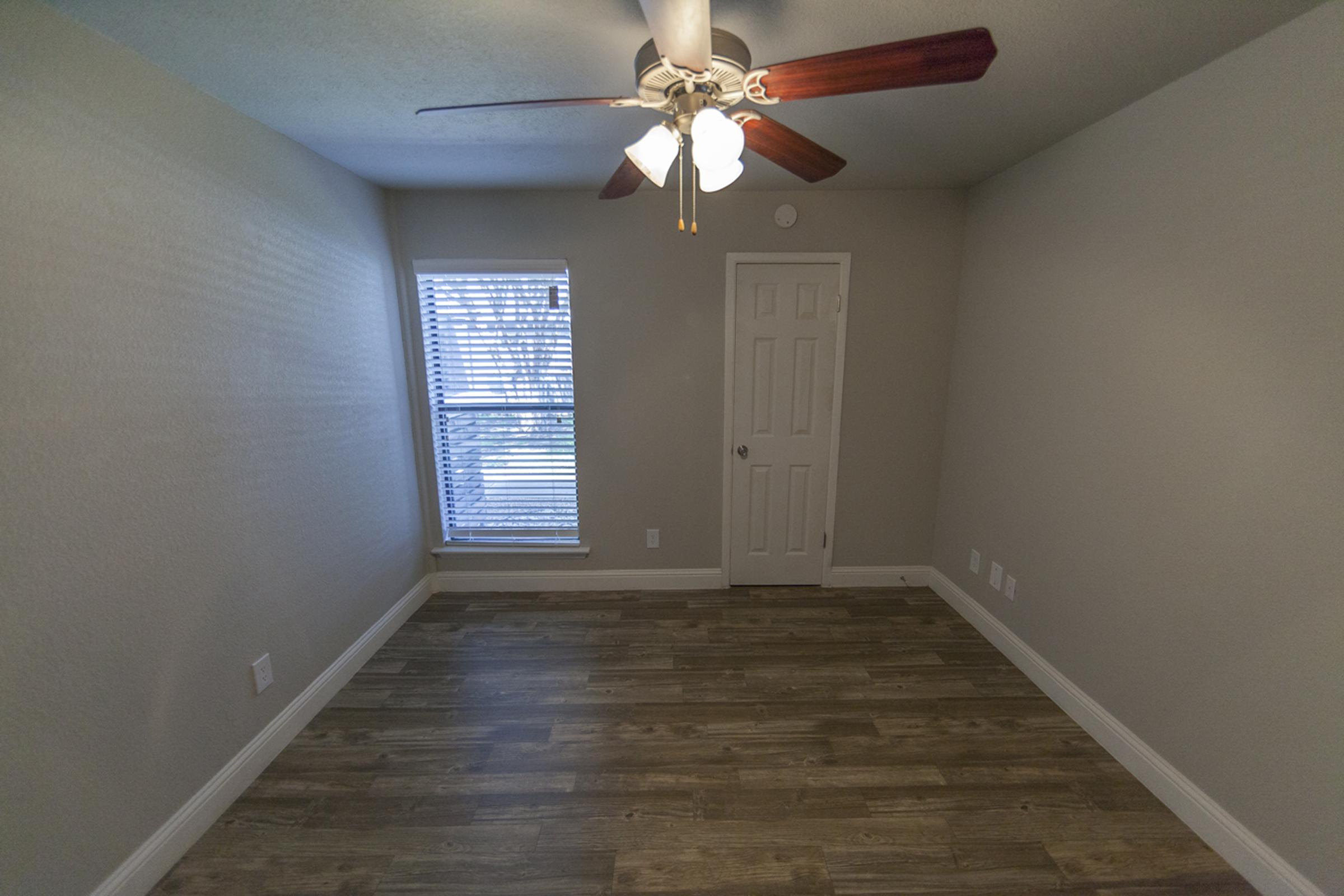 A small, empty room with a ceiling fan and hardwood-style flooring. A window with blinds allows natural light to enter, illuminating the light gray walls. The room features a single door and white trim, creating a clean and airy atmosphere.