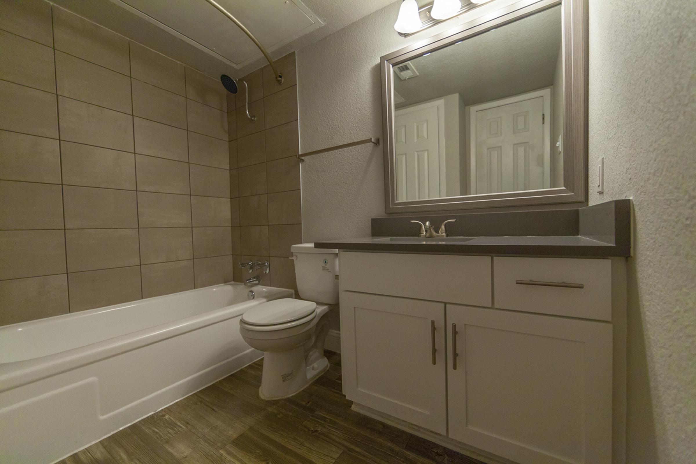 Modern bathroom featuring a white bathtub, toilet, and a single sink vanity with a dark countertop. The walls are adorned with beige tiles, and there's a large mirror above the sink. Soft lighting is provided by fixtures above the mirror, and the floor has a wood-like finish.